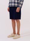 A model wears the Indigo Recycled Herringbone Pleated Track Short against a light grey background. The image shows the model from the waist down, highlighting the shorts' relaxed fit, herringbone texture, and pleated front. The model's hands are placed in the side pockets, and the shorts are paired with a dark top.