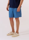 A model wears the Washed Indigo Recycled Herringbone Pleated Track Short against a plain white background. The shorts feature prominent front pleats, a relaxed fit, and a visible herringbone texture in a washed indigo blue shade. The image highlights the garment’s construction from the waist down, showing the elasticated waistband and side pockets.