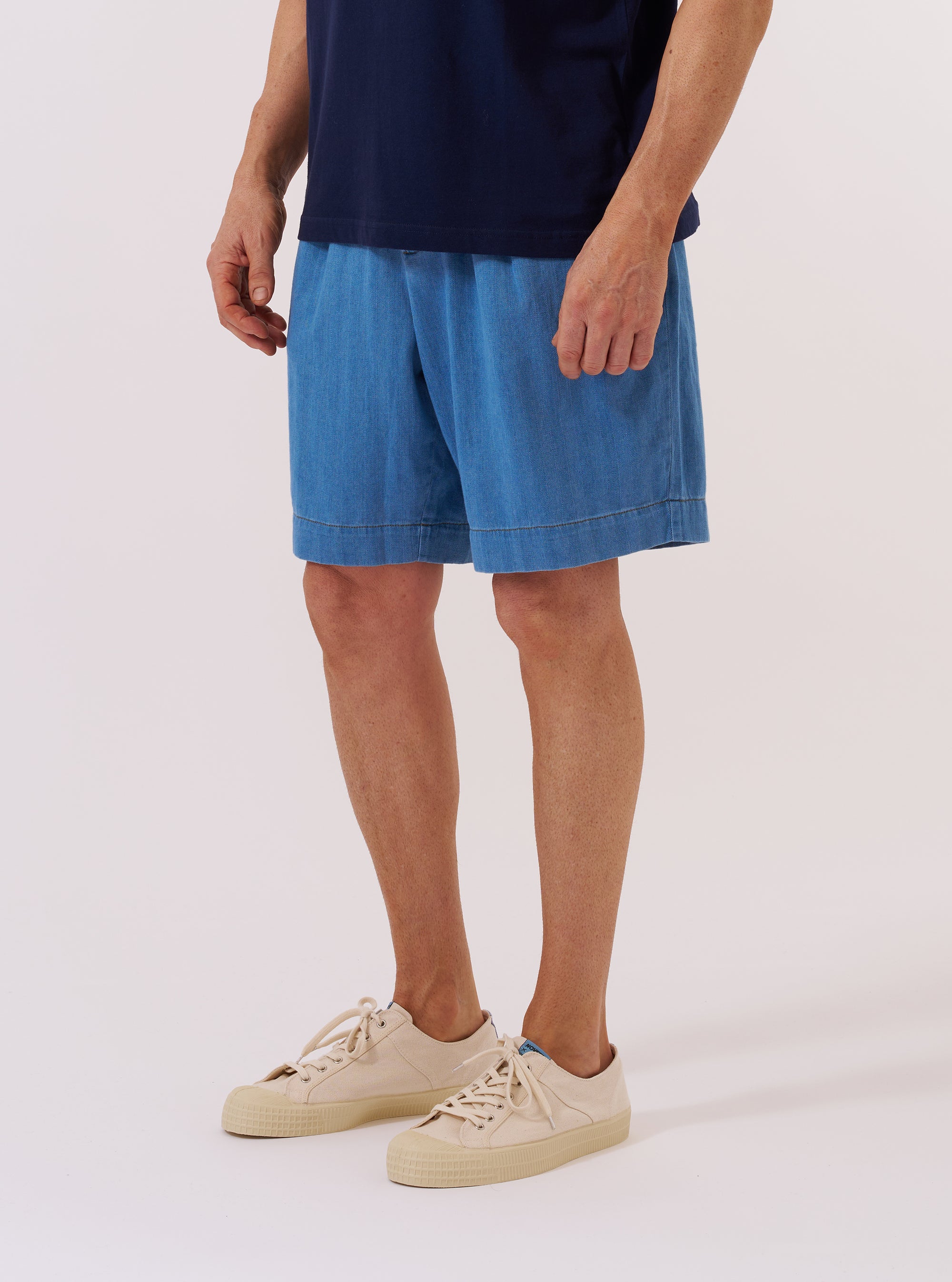A model wears the Washed Indigo Recycled Herringbone Pleated Track Short against a plain white background. The shorts feature prominent front pleats, a relaxed fit, and a visible herringbone texture in a washed indigo blue shade. The image highlights the garment’s construction from the waist down, showing the elasticated waistband and side pockets.