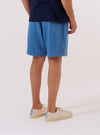 A model is wearing the Washed Indigo Recycled Herringbone Pleated Track Short, shown from a rear three-quarter angle against a plain white background. The shorts feature a relaxed fit with an elasticated waistband and a single rear patch pocket. The recycled cotton fabric is a washed indigo blue and displays a distinct herringbone texture. No mannequin is present in the image.