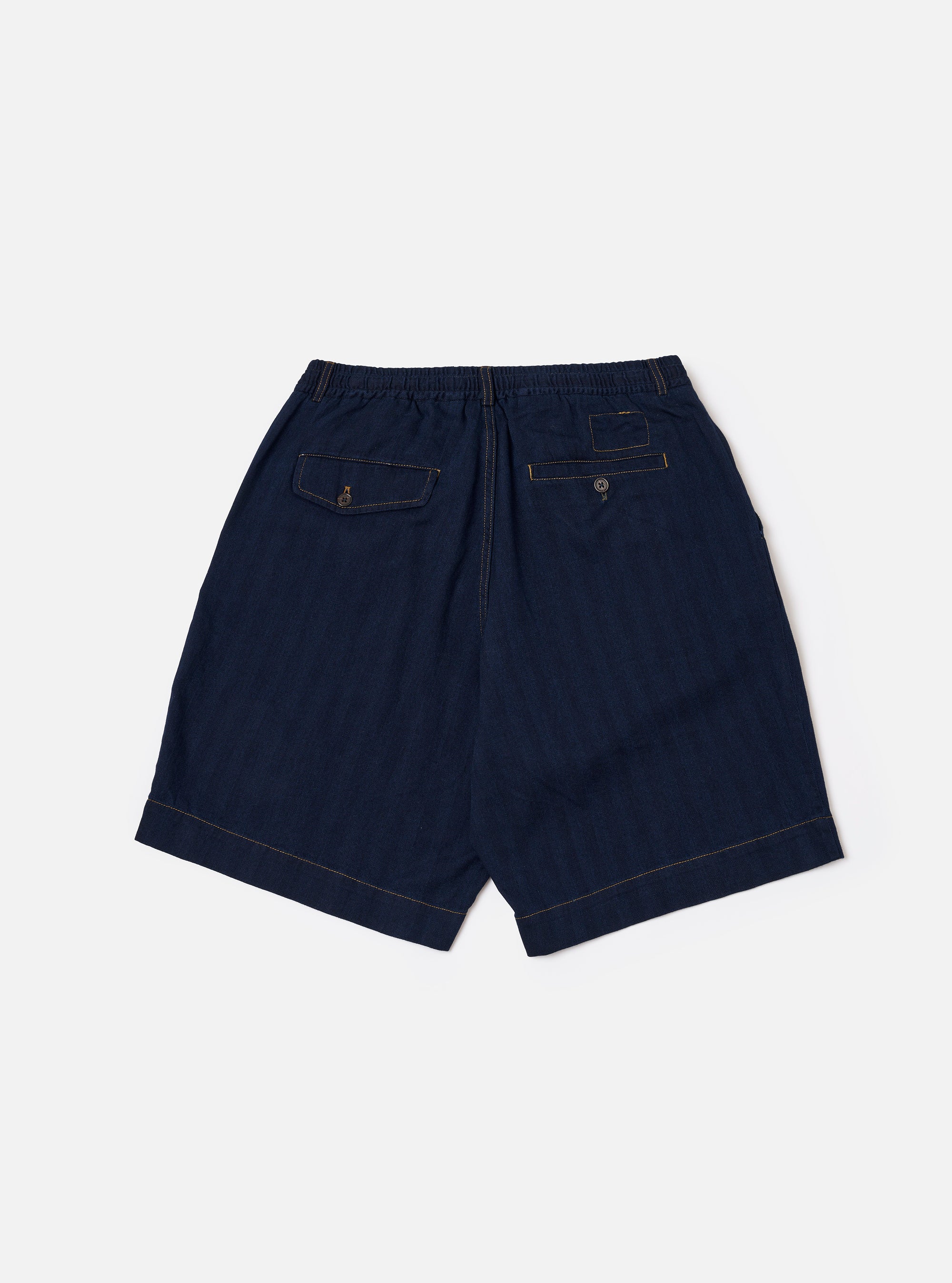 A model wears the Indigo Recycled Herringbone Pleated Track Short, shown from the waist down against a plain white background. The shorts feature a relaxed fit with a single pleat at the front and side pockets. The indigo-dyed fabric has a visible herringbone texture.
