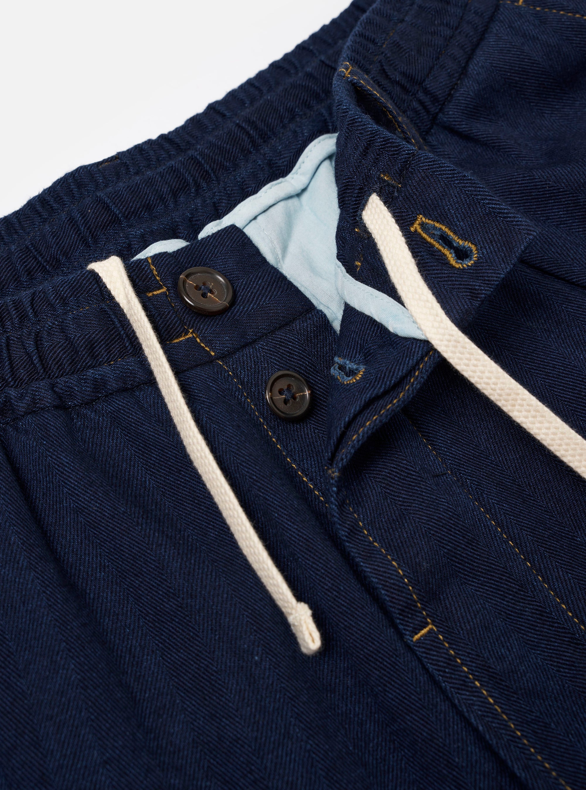 A model is wearing the Indigo Recycled Herringbone Pleated Track Short, shown from the rear against a plain white background. The shorts feature a visible indigo-colored herringbone weave, an elasticated waistband, and two rear pockets: one with a buttoned flap and one jetted pocket with a button closure.