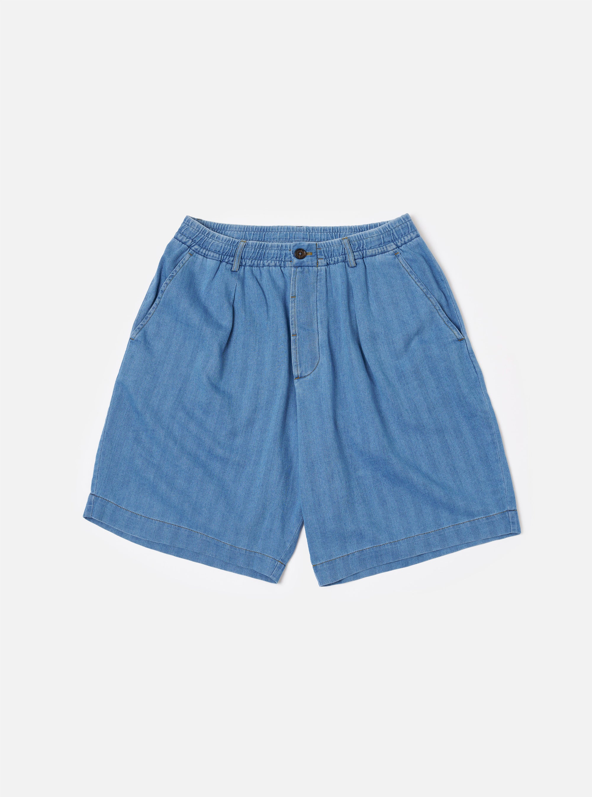 The Washed Indigo Recycled Herringbone Pleated Track Short is displayed as a flat lay against a white background. The shorts feature a relaxed fit with an elasticated waistband, an internal drawstring, a single front pleat on each leg, and two side pockets. The fabric is a washed indigo blue recycled cotton with a visible herringbone texture.