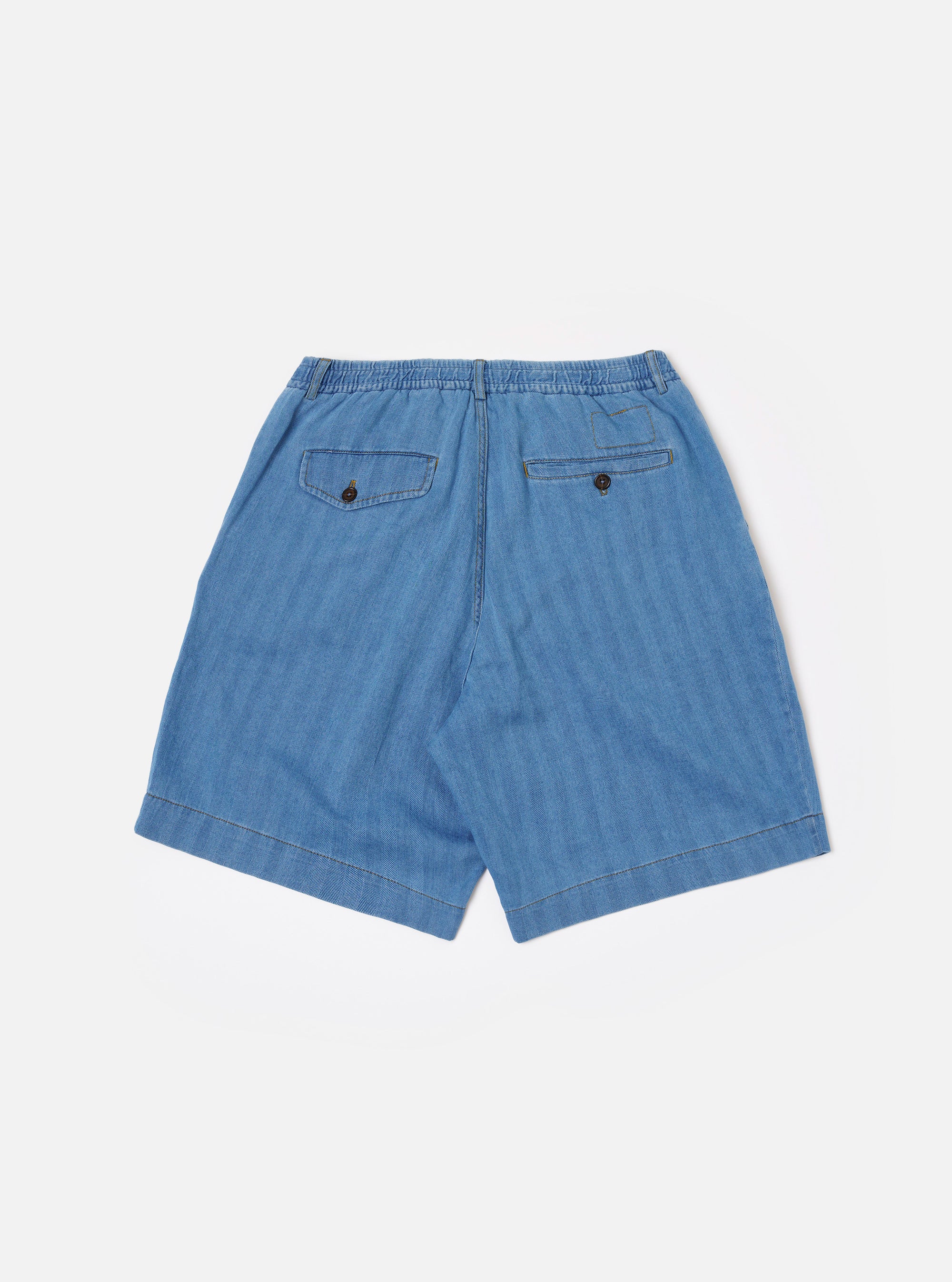 The Washed Indigo Recycled Herringbone Pleated Track Short is shown from the back as a flat lay against a plain white background. The image highlights the rear construction, featuring one buttoned flap pocket on the left and one buttoned jetted pocket on the right. The elasticated waistband and the textured herringbone weave of the washed indigo fabric are clearly visible.