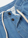 The Washed Indigo Recycled Herringbone Pleated Track Short is shown from a rear angle as a flat lay against a plain white background. The image highlights the elasticated waistband and two rear pockets, including one with a buttoned flap and one jetted pocket. The washed indigo blue fabric features a distinct herringbone weave.