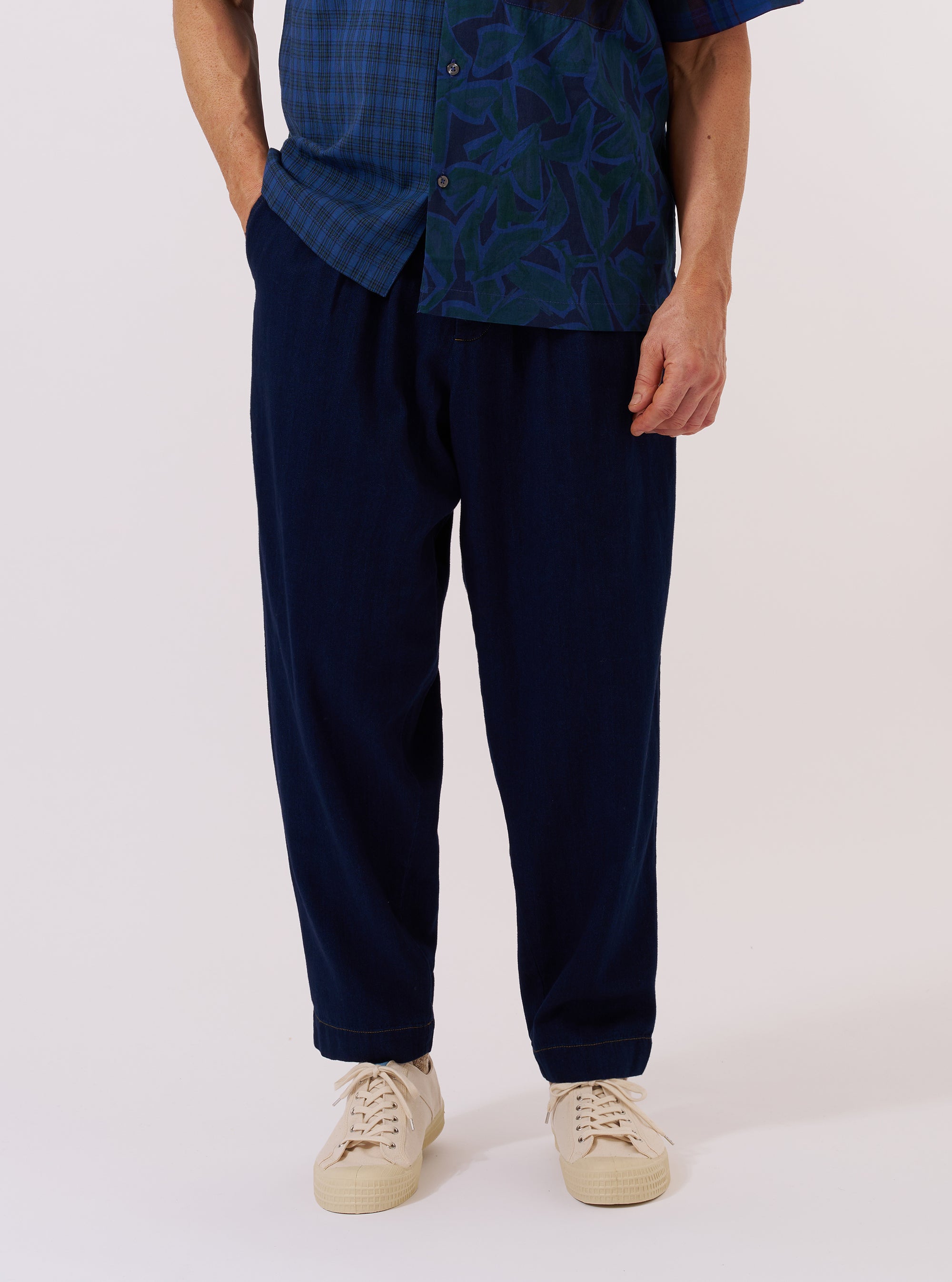 A model wears the Indigo Recycled Herringbone Pleated Track Pant against a plain white background. The image shows a front view of the trousers, highlighting the relaxed, tapered fit, the elasticated drawstring waistband, and the single pleat at the front of each leg. The fabric is a deep indigo blue with a visible herringbone weave. The model is wearing the pants with a tucked-in shirt and dark shoes.