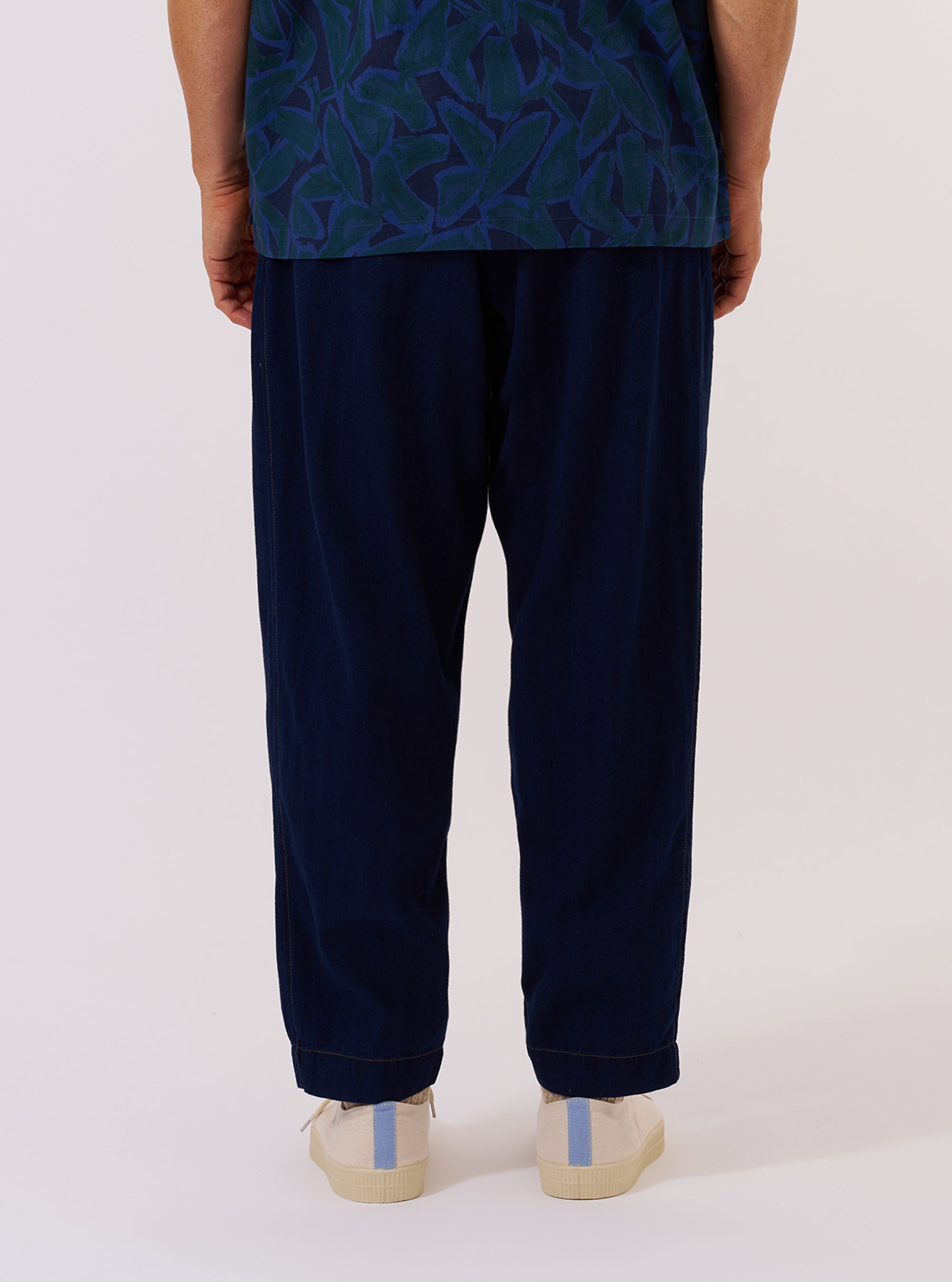 A model wears the Indigo Recycled Herringbone Pleated Track Pant against a plain white background. The image shows the model from the waist down, highlighting the loose, tapered fit and the single pleat at the front of each leg. The dark indigo fabric features a distinct herringbone weave. The model is wearing the trousers with white socks and dark footwear.