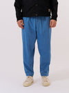 A model wears the Washed Indigo Recycled Herringbone Pleated Track Pant while standing against a plain white background. The trousers feature a loose, tapered fit with a pleated front and an elasticated drawstring waist. The garment is made from a washed indigo blue fabric with a visible herringbone weave.