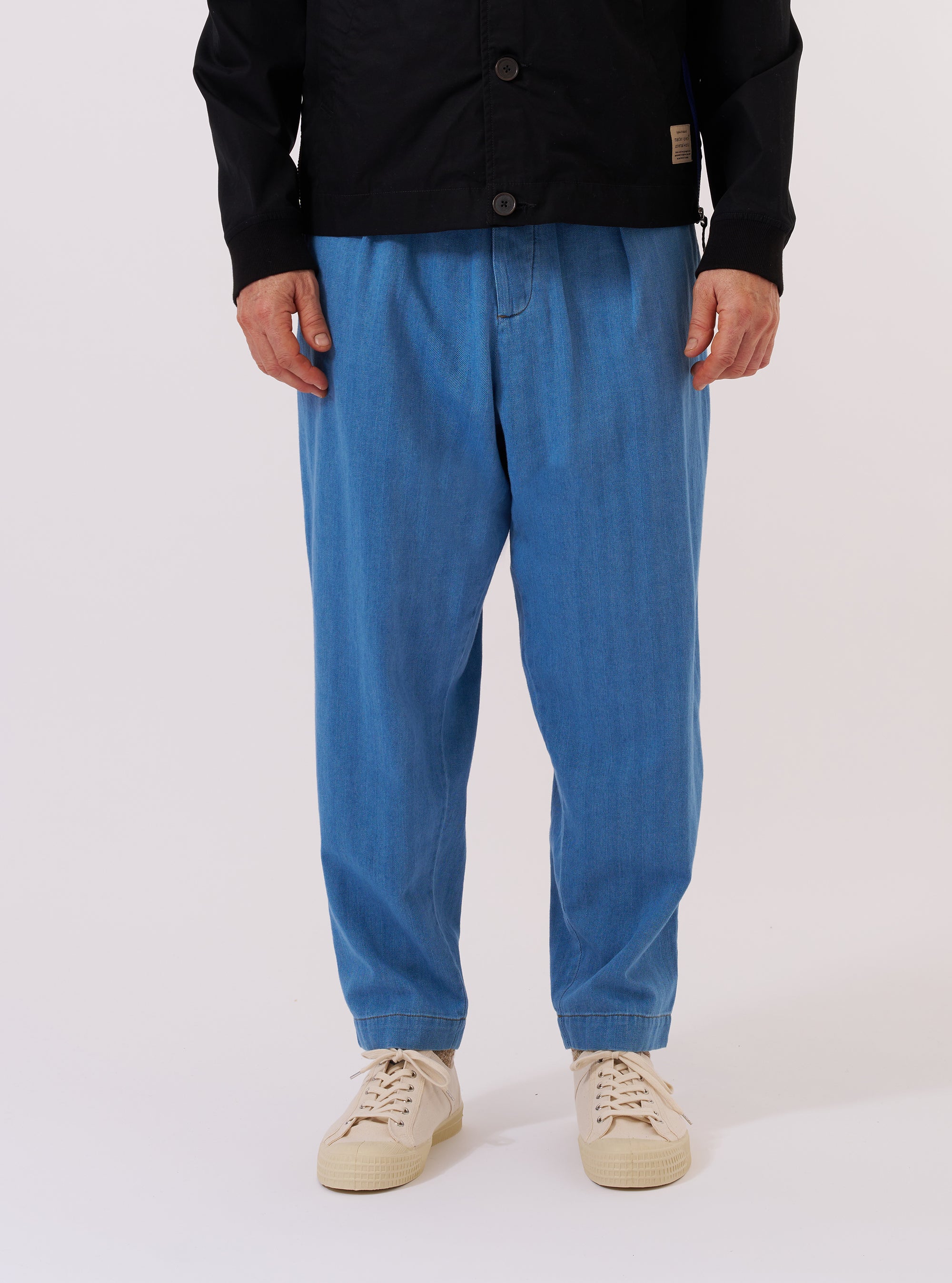 A model wears the Washed Indigo Recycled Herringbone Pleated Track Pant while standing against a plain white background. The trousers feature a loose, tapered fit with a pleated front and an elasticated drawstring waist. The garment is made from a washed indigo blue fabric with a visible herringbone weave.