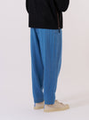 A model wears the Washed Indigo Recycled Herringbone Pleated Track Pant, shown from the rear three-quarter angle against a plain white background. The image highlights the loose, tapered fit and the textured herringbone weave of the washed indigo fabric. One rear patch pocket is visible on the right side, and the elasticated waistband is prominent. The model is also wearing a light-colored long-sleeve shirt and dark shoes.