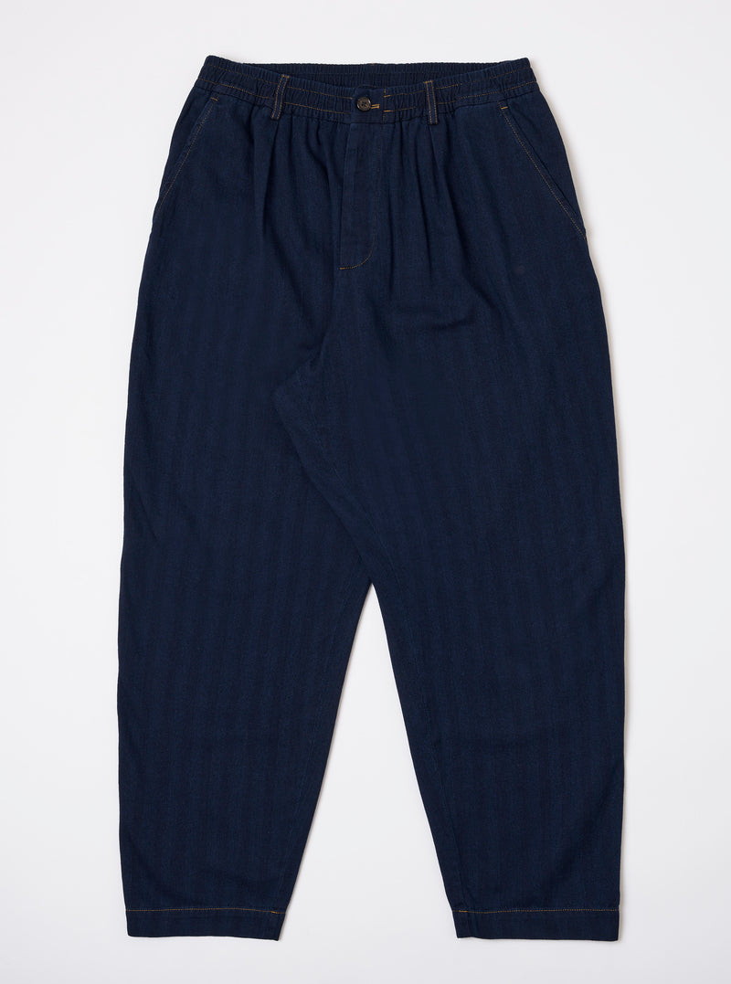 The Indigo Recycled Herringbone Pleated Track Pant is shown as a flat lay against a plain white background. The trousers feature a drawstring waistband, a single pleat on each leg, and a relaxed, tapered fit. The dark indigo fabric has a visible herringbone texture.