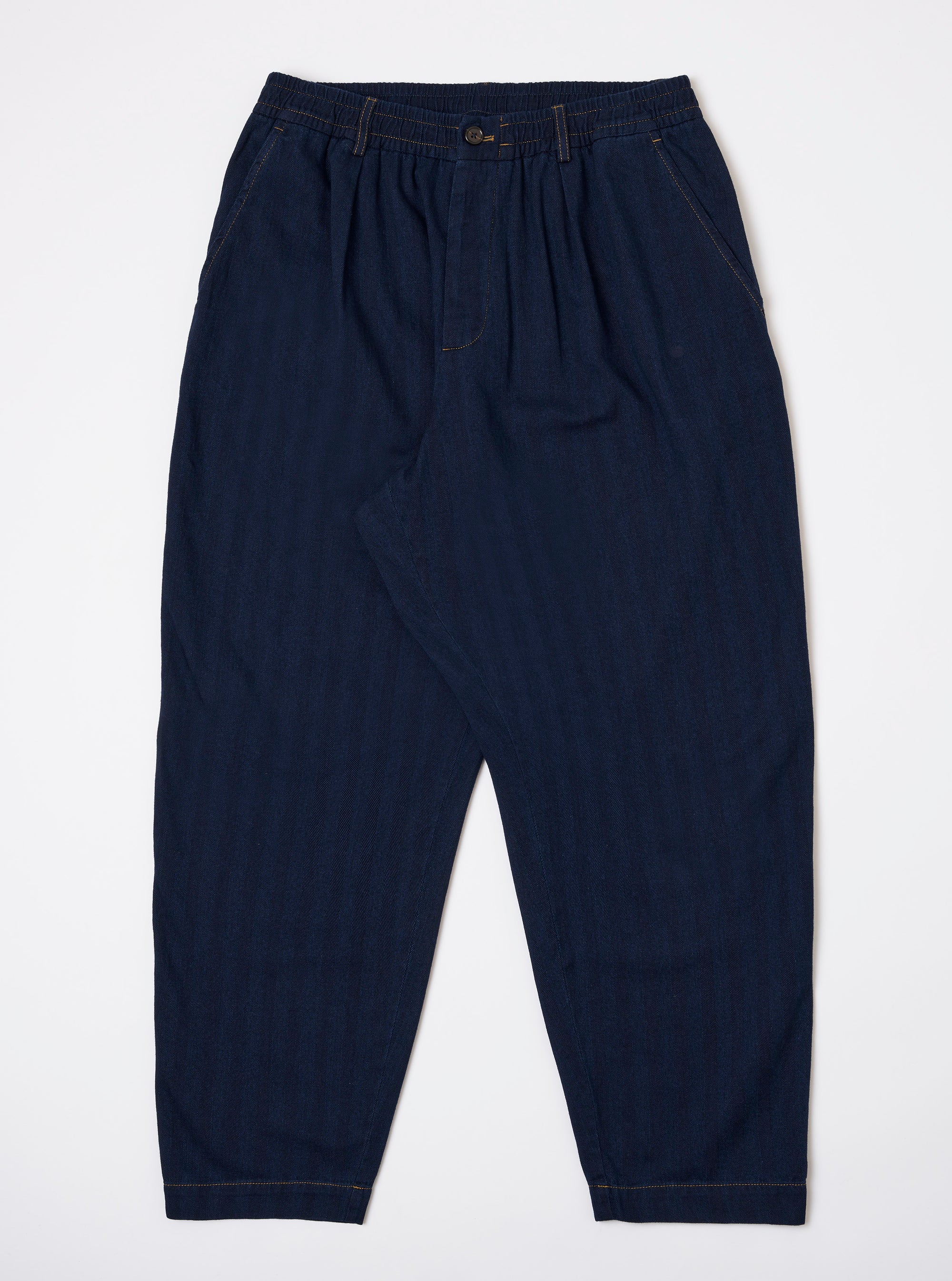 The Indigo Recycled Herringbone Pleated Track Pant is shown as a flat lay against a plain white background. The trousers feature a drawstring waistband, a single pleat on each leg, and a relaxed, tapered fit. The dark indigo fabric has a visible herringbone texture.