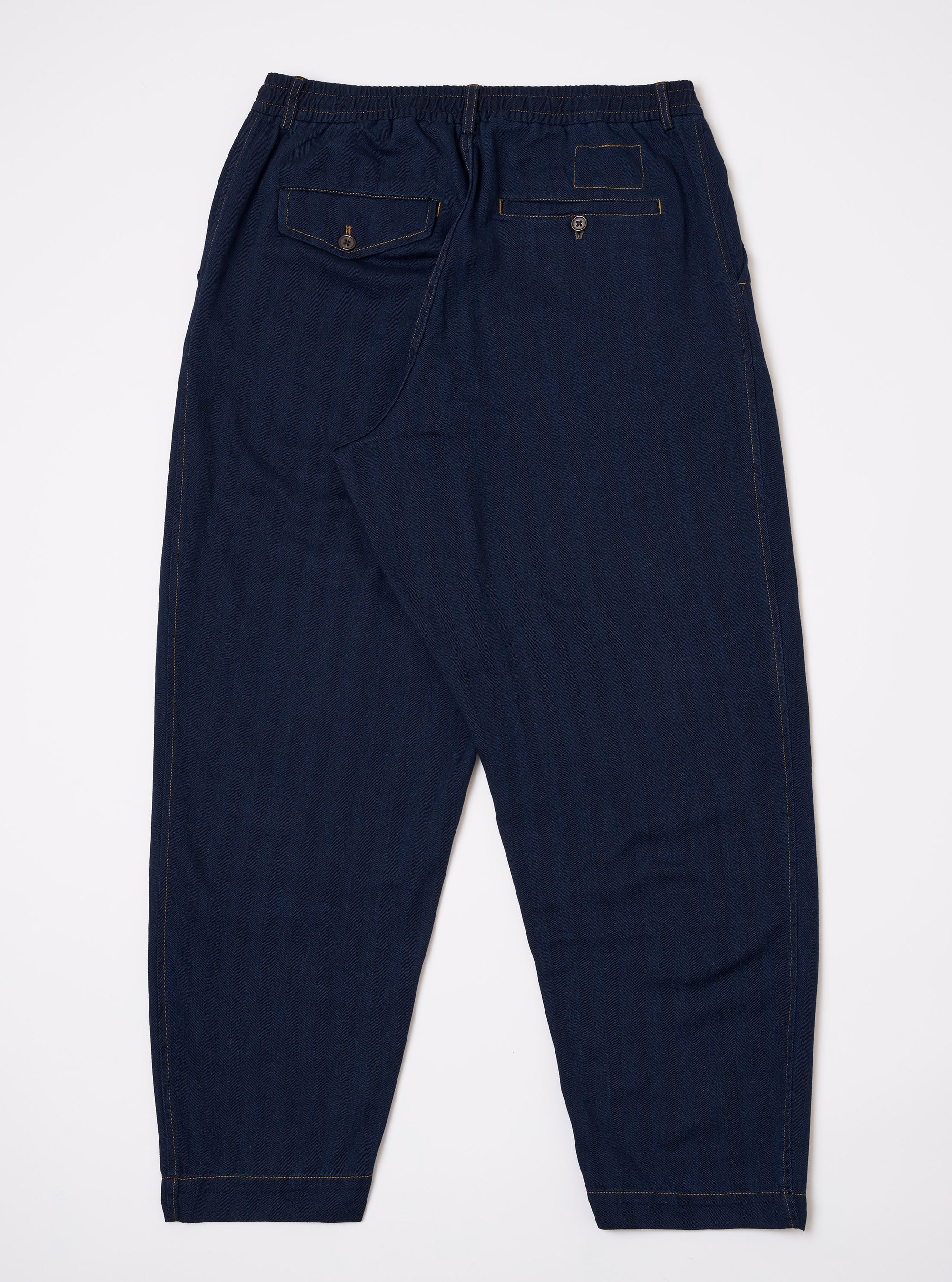 The Indigo Recycled Herringbone Pleated Track Pant is shown from the back as a flat lay against a plain white background. The image highlights the elasticated waistband and the two rear patch pockets. The indigo-colored fabric features a visible herringbone weave and tonal stitching.