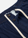 The Indigo Recycled Herringbone Pleated Track Pant is shown from the back as a flat lay against a plain white background. The image highlights the two rear pockets, including one with a buttoned flap and one buttoned jetted pocket, as well as the elasticated waistband and the textured herringbone weave of the indigo blue fabric.