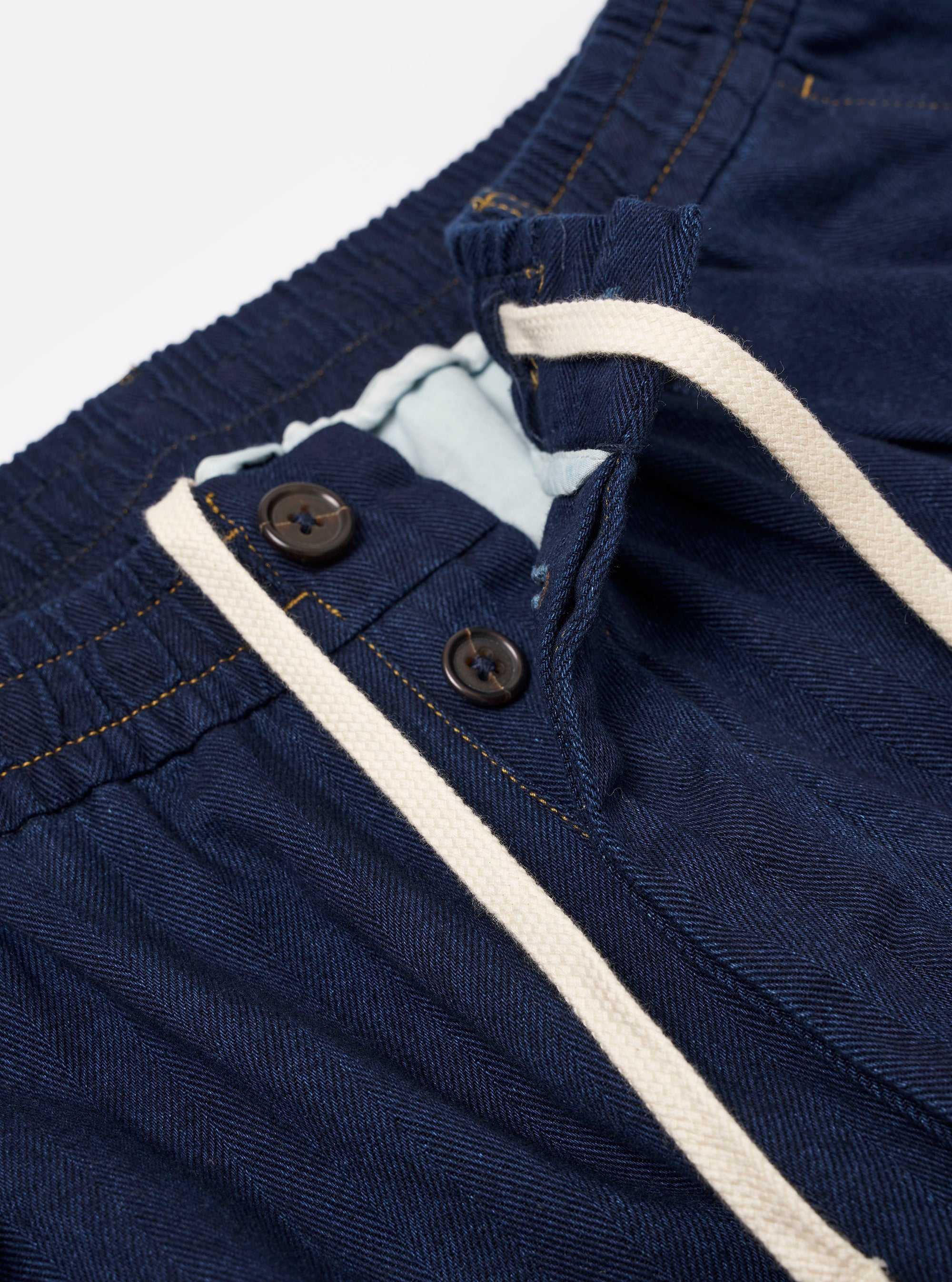 The Indigo Recycled Herringbone Pleated Track Pant is shown from the back as a flat lay against a plain white background. The image highlights the two rear pockets, including one with a buttoned flap and one buttoned jetted pocket, as well as the elasticated waistband and the textured herringbone weave of the indigo blue fabric.
