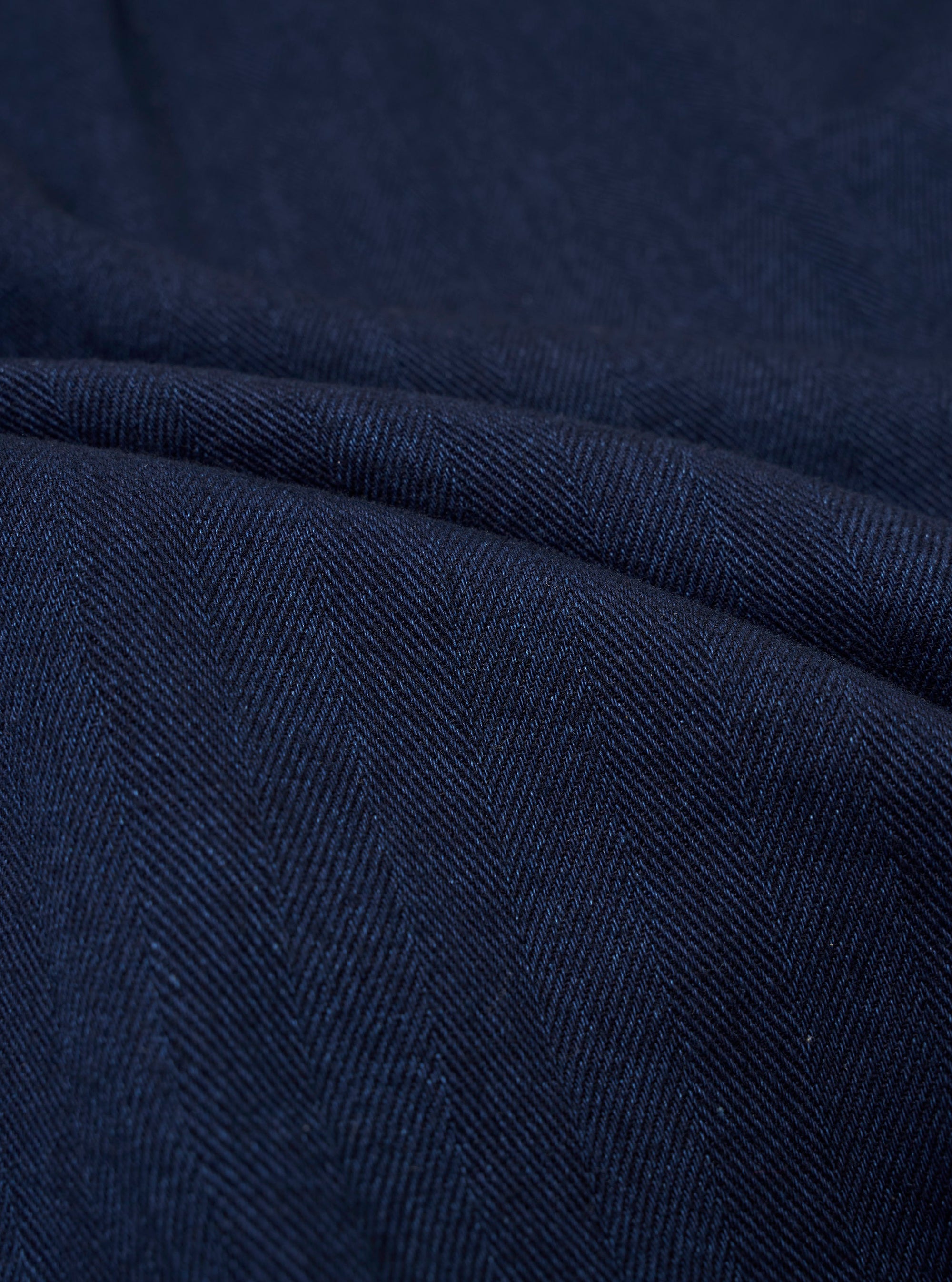 A close-up swatch image of the Indigo Recycled Herringbone Pleated Track Pant fabric highlights the intricate V-shaped herringbone weave. The textile features a deep indigo blue color with a textured, three-dimensional appearance.