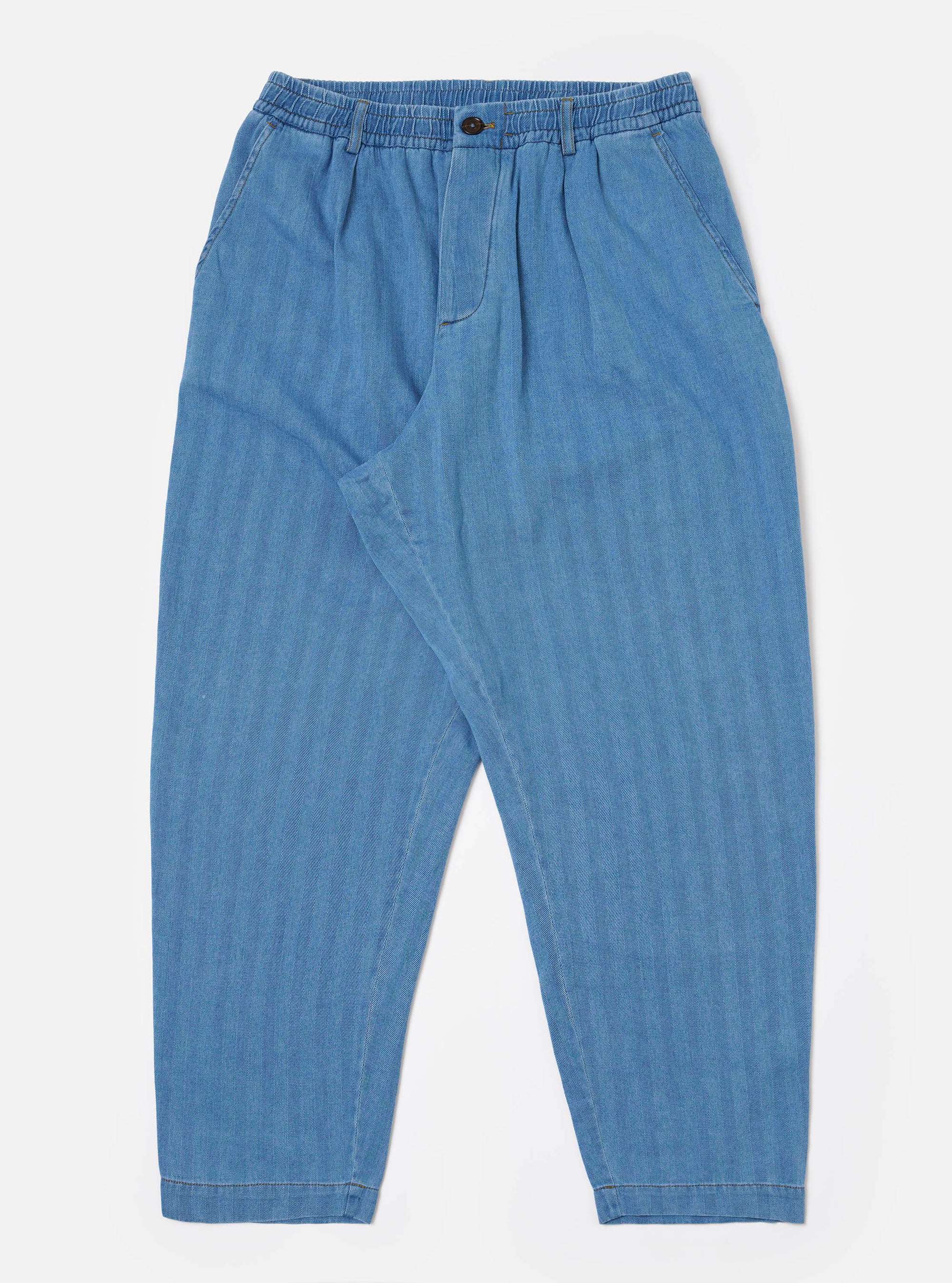 The Washed Indigo Recycled Herringbone Pleated Track Pant is shown as a flat lay against a white background. The trousers feature an elasticated waistband with an external drawstring, a single pleat at the front, and a loose, tapered leg. Two side pockets are visible, and the fabric displays a distinct herringbone texture in a washed indigo blue.