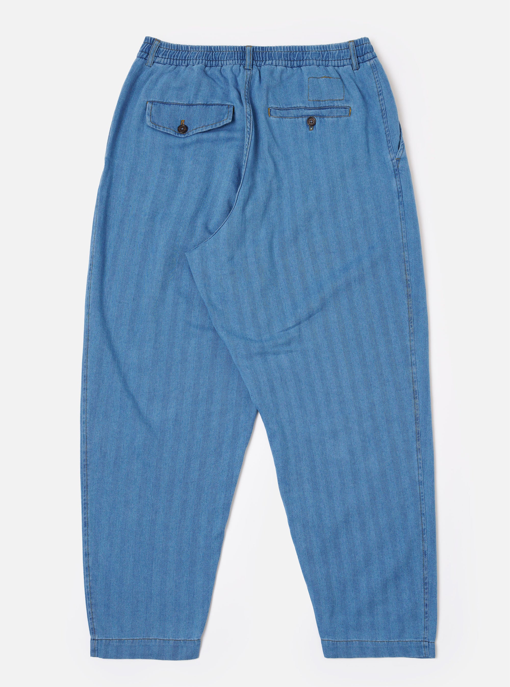 The Washed Indigo Recycled Herringbone Pleated Track Pant is shown from the back as a flat lay against a plain white background. The image highlights the elasticated waistband and the two rear pockets, one of which features a buttoned flap. The washed indigo fabric has a visible herringbone texture.