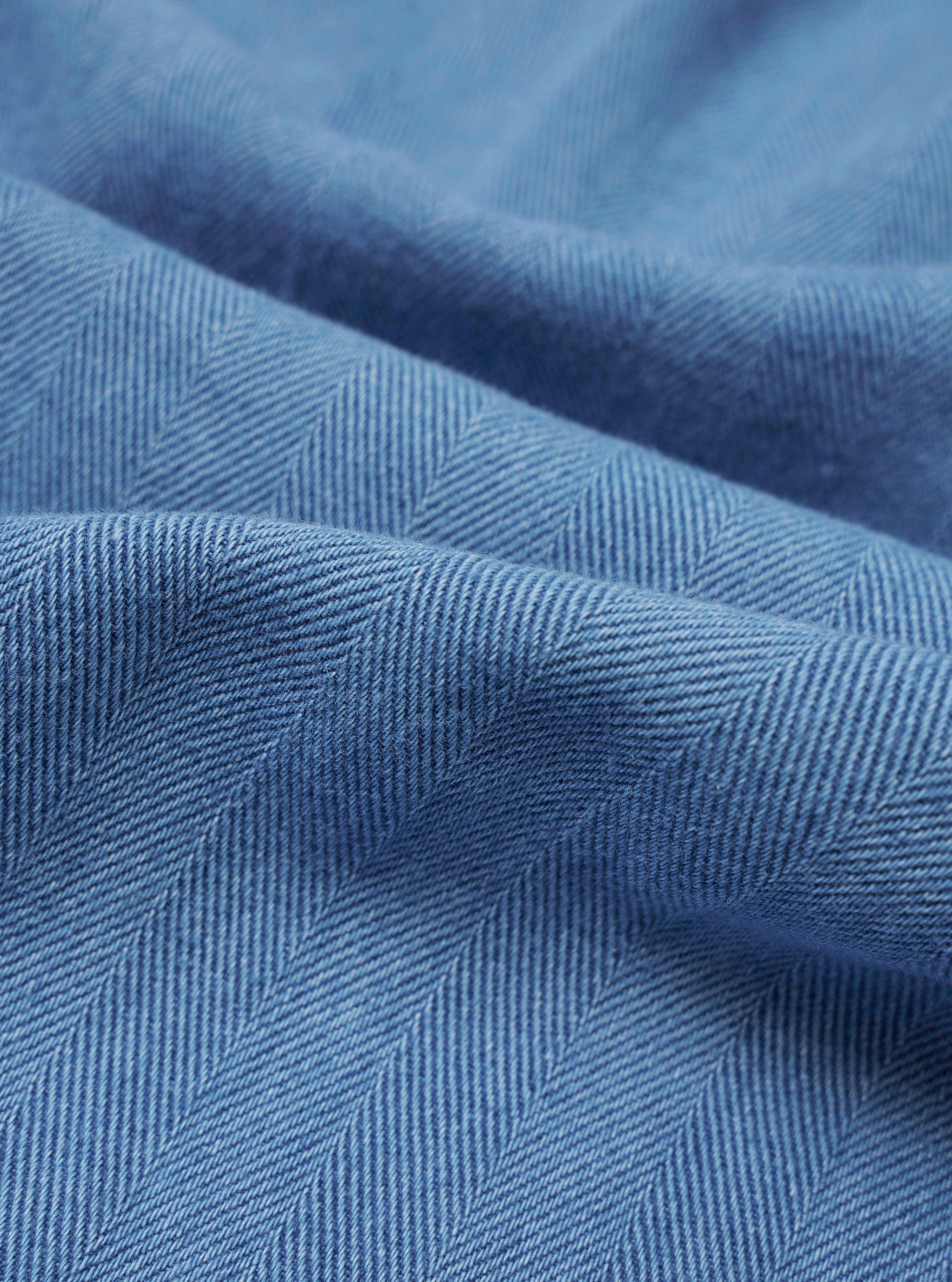 A close-up swatch image of the Washed Indigo Recycled Herringbone Pleated Track Pant fabric highlights the blue washed indigo color and the distinct V-shaped herringbone weave. The textured recycled cotton material fills the frame, providing a detailed view of the garment's textile construction.