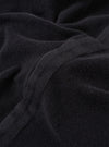 The Black Light Weight Terry Newlyn Polo is shown in a detail view as a flat lay against a plain white background. The image highlights the soft, looped texture of the black light weight terry fabric and the clean construction of the garment's hem and side split.