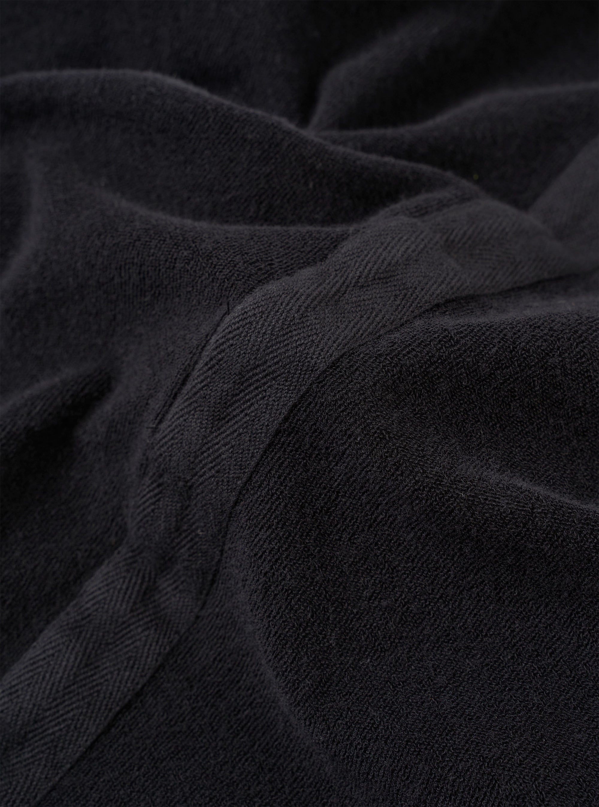 The Black Light Weight Terry Newlyn Polo is shown in a detail view as a flat lay against a plain white background. The image highlights the soft, looped texture of the black light weight terry fabric and the clean construction of the garment's hem and side split.