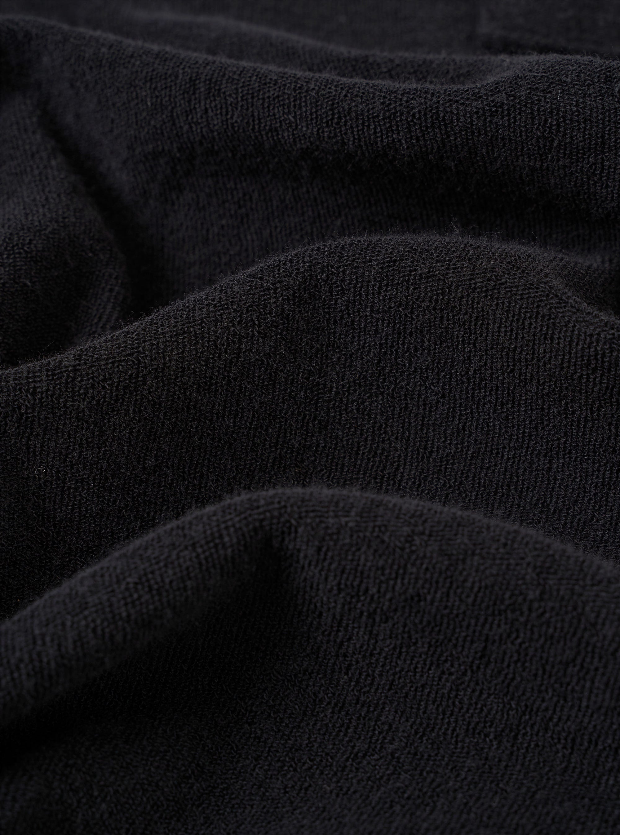 A close-up fabric swatch of the Black Light Weight Terry Newlyn Polo highlights the plush, looped texture of the fine terry cloth. The image focuses on the deep black color and the soft, technical detail of the material.