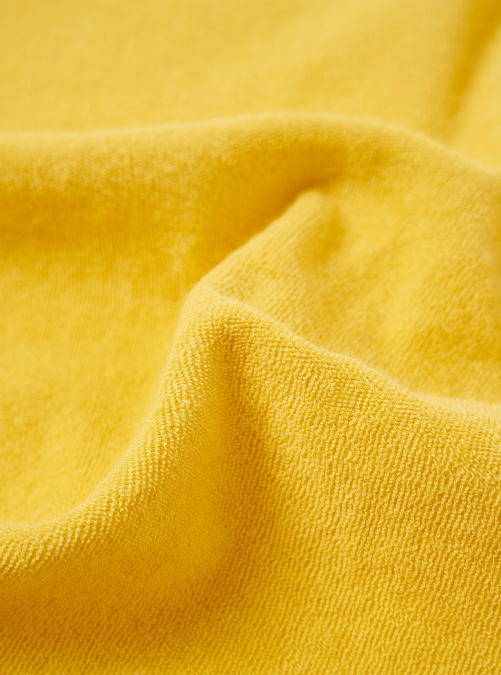A close-up fabric swatch of the Yellow Light Weight Terry Newlyn Polo highlights the soft, looped texture of the yellow cotton terry material. The fabric fills the entire frame, providing a detailed view of the garment's color and knit construction.