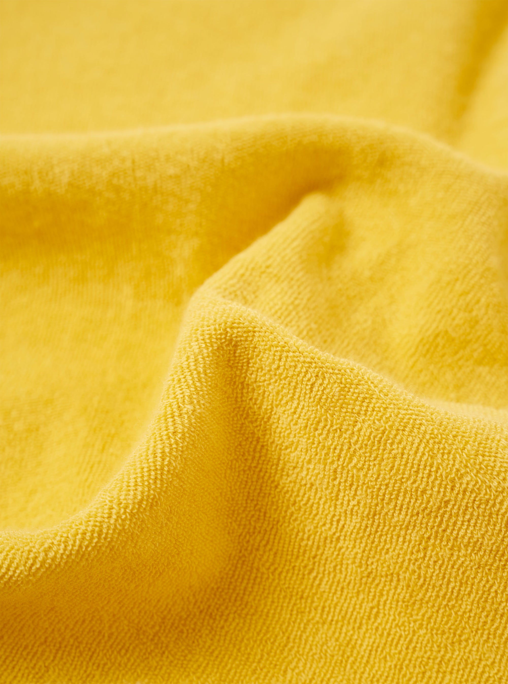 A close-up fabric swatch of the Yellow Light Weight Terry Newlyn Polo highlights the soft, looped texture of the yellow cotton terry material. The fabric fills the entire frame, providing a detailed view of the garment's color and knit construction.