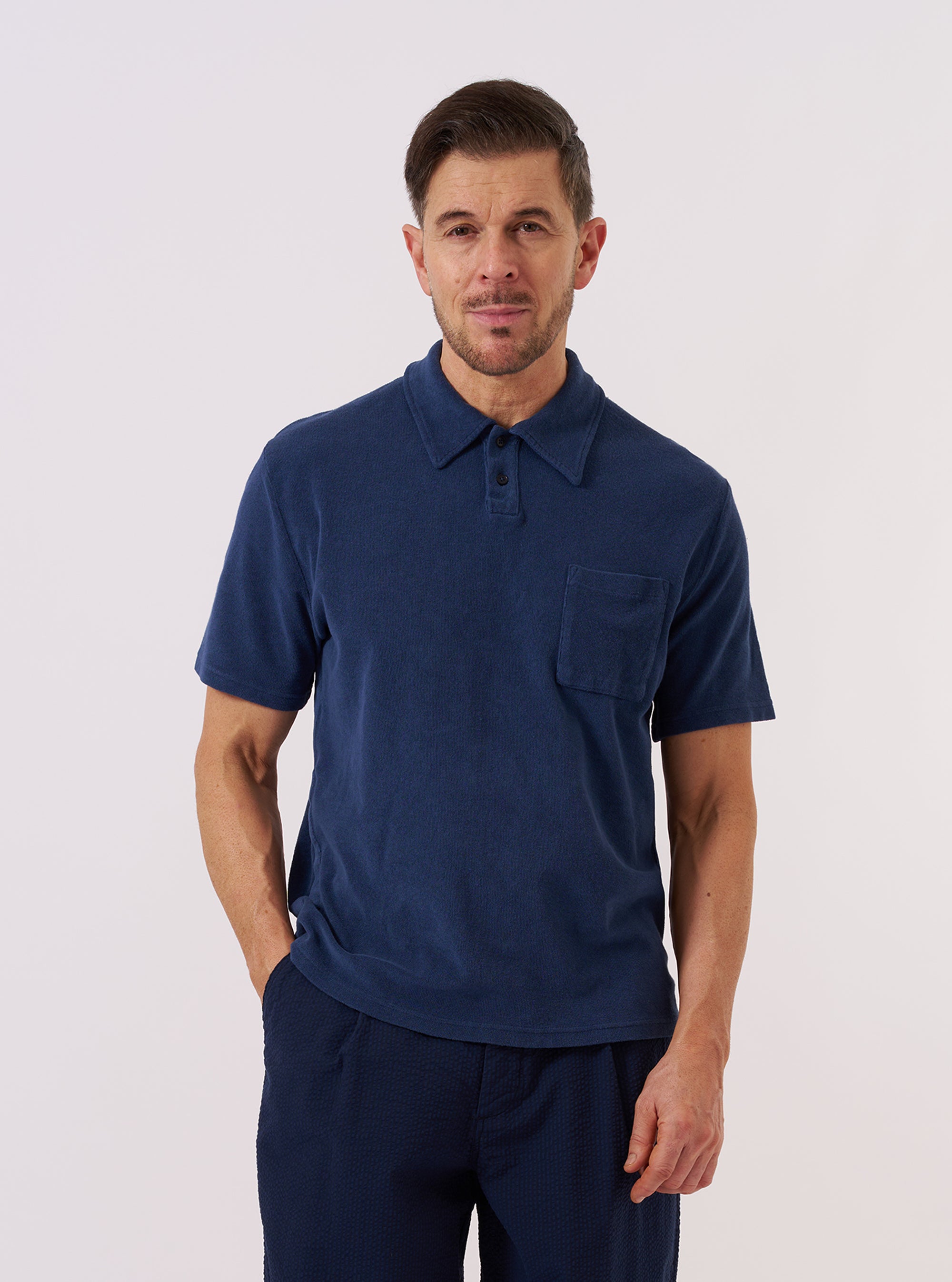A model wears the Navy Light Weight Terry Newlyn Polo against a plain white background. The polo features a relaxed fit, a self-fabric one-piece collar, a two-button placket, and a large patch pocket on the left chest. The navy blue fabric has a soft, looped terry texture. The model is shown from the front, highlighting the garment's simple, overhead construction and split hem.