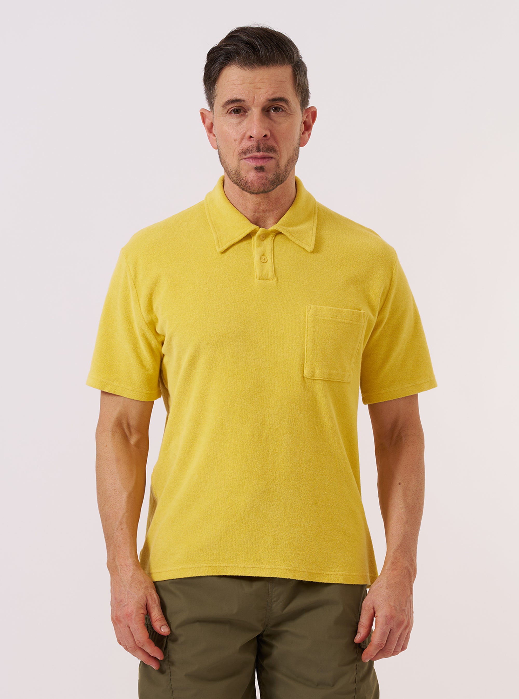 A model wears the Yellow Light Weight Terry Newlyn Polo while standing against a plain white background. The polo features a classic collar, a short button placket, and short sleeves. The garment is made from a vibrant yellow light weight terry fabric with a soft, textured appearance.