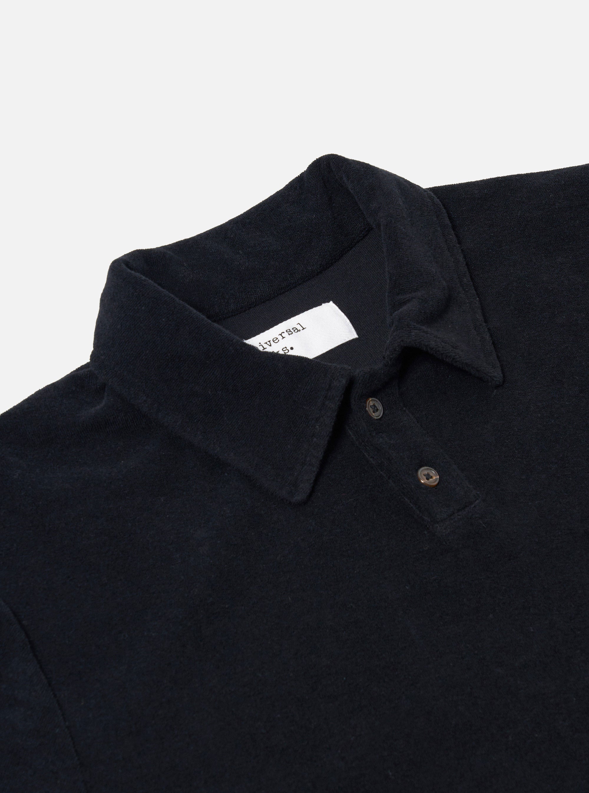 The Black Light Weight Terry Newlyn Polo is shown as a flat lay against a plain white background. The polo features a classic collar, a three-button placket, and short sleeves. The black cotton fabric has a visible light-weight terry texture, and the garment is finished with a straight hem.