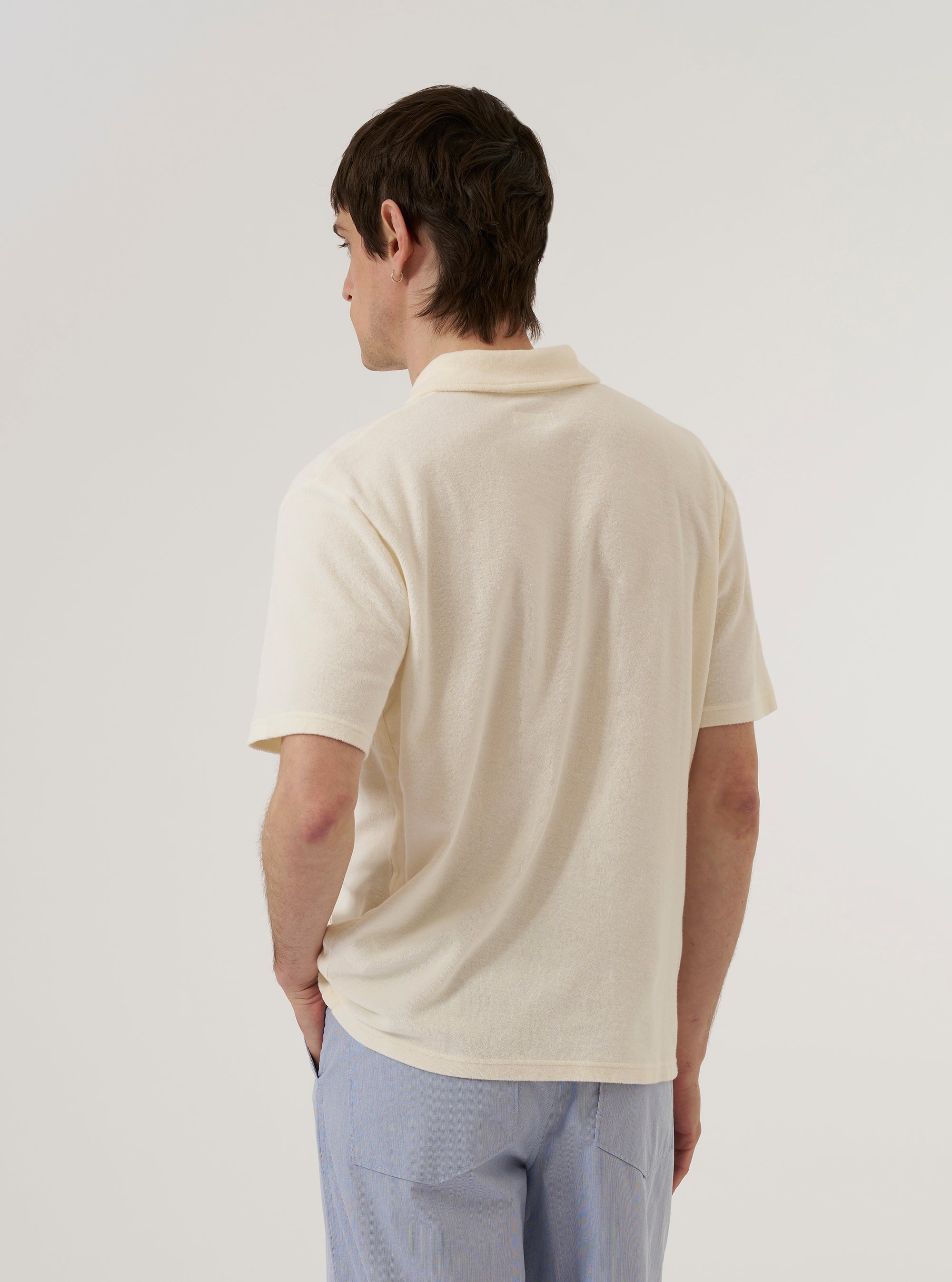 A model wears the Ecru Light Weight Terry Newlyn Polo against a plain white background, shown from a rear three-quarter angle. The polo features a relaxed fit, short sleeves, and a simple straight hem. The light-colored terry fabric has a soft, textured appearance.