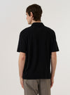 A model wears the Black Light Weight Terry Newlyn Polo, shown from the back against a plain white background. The polo is made from a soft, black lightweight terry cotton and features short sleeves, a structured collar, and a straight hem. The image highlights the relaxed fit and the textured finish of the fabric.