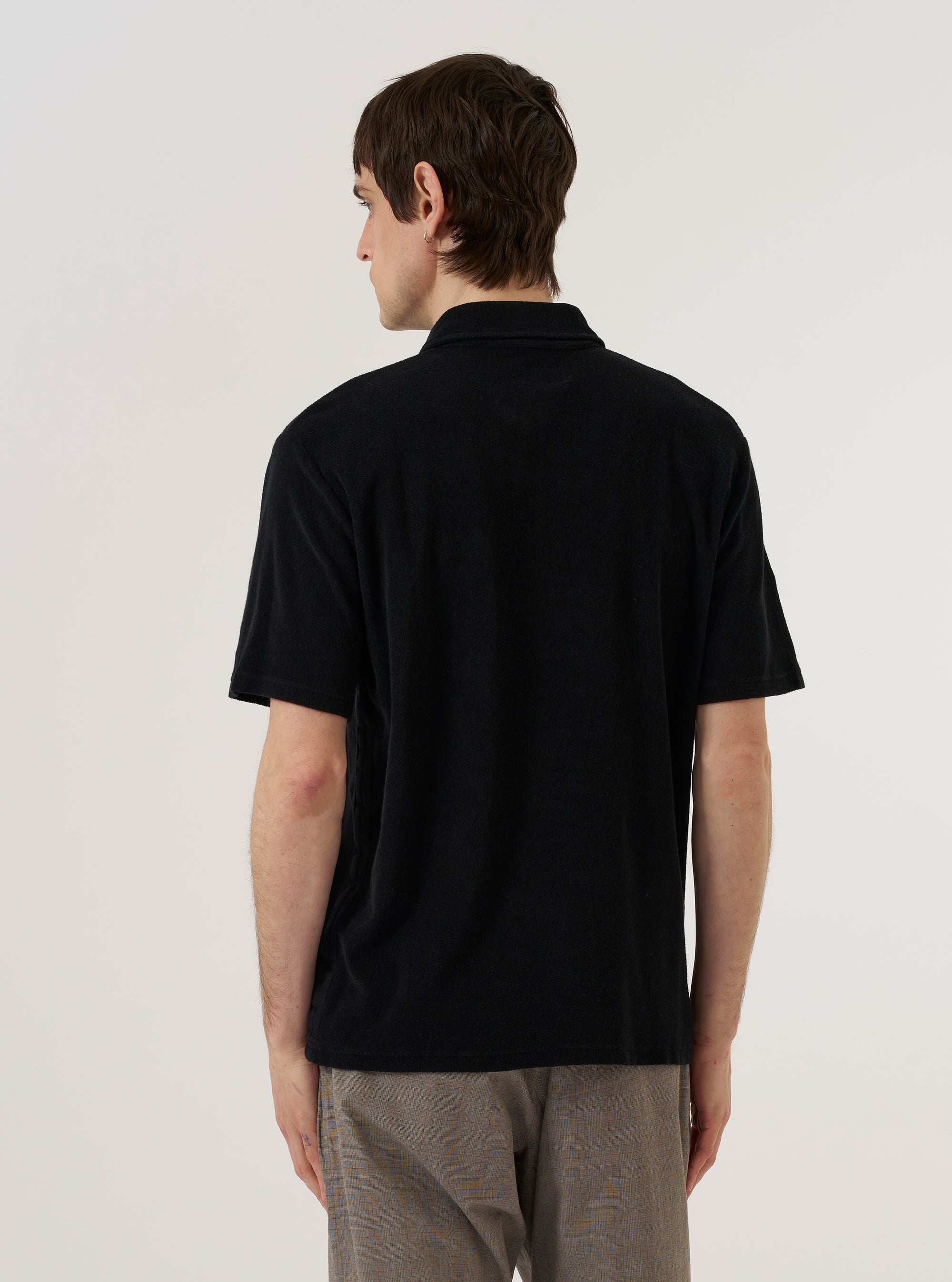 A model wears the Black Light Weight Terry Newlyn Polo, shown from the back against a plain white background. The polo is made from a soft, black lightweight terry cotton and features short sleeves, a structured collar, and a straight hem. The image highlights the relaxed fit and the textured finish of the fabric.
