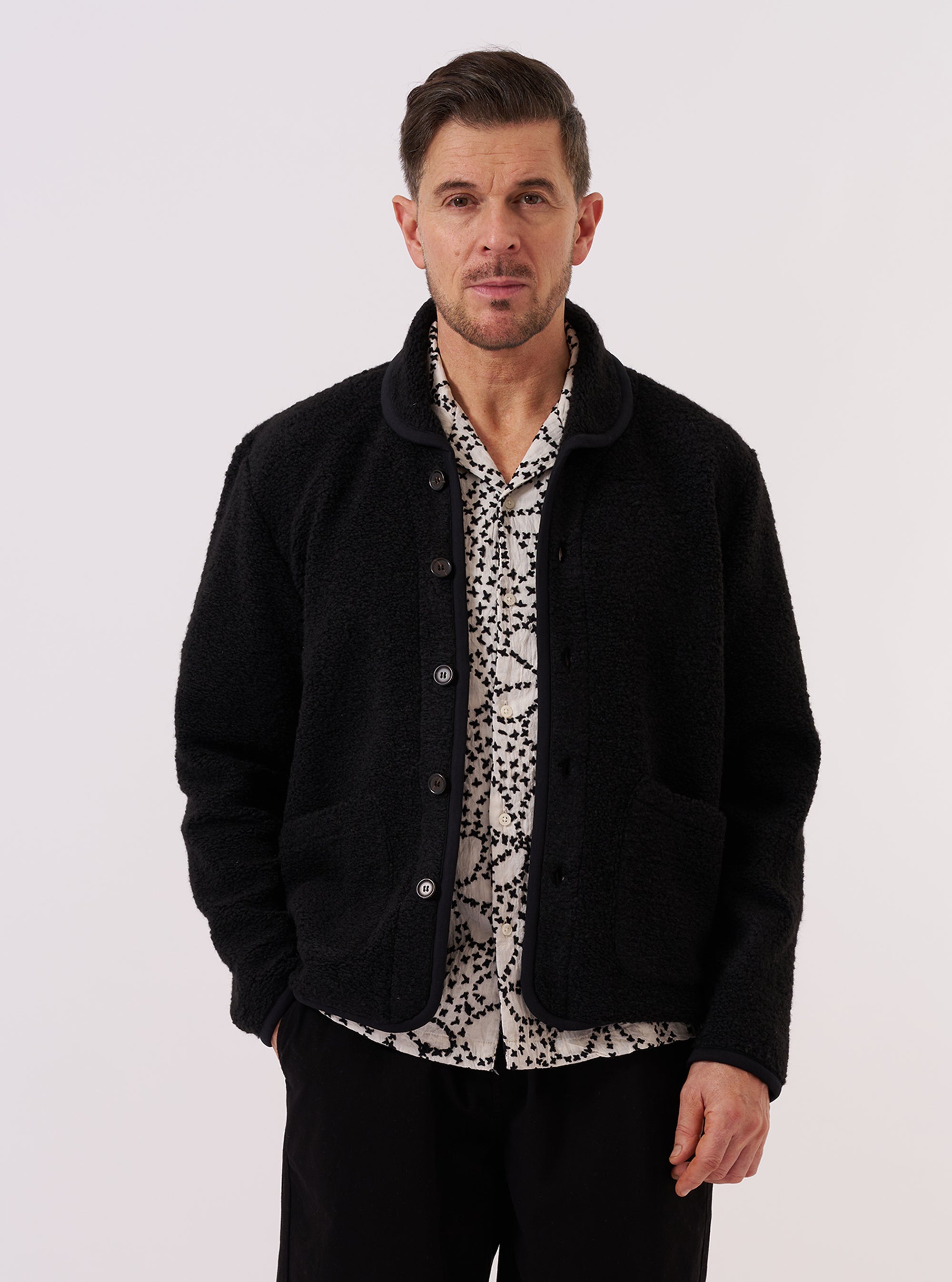 A model wears the Black Alvar Fleece Lancaster Jacket while standing against a plain white background. The jacket features a rounded shawl collar, a six-button front closure, and two large patch pockets at the waist. The black fabric has a soft, textured fleece appearance.