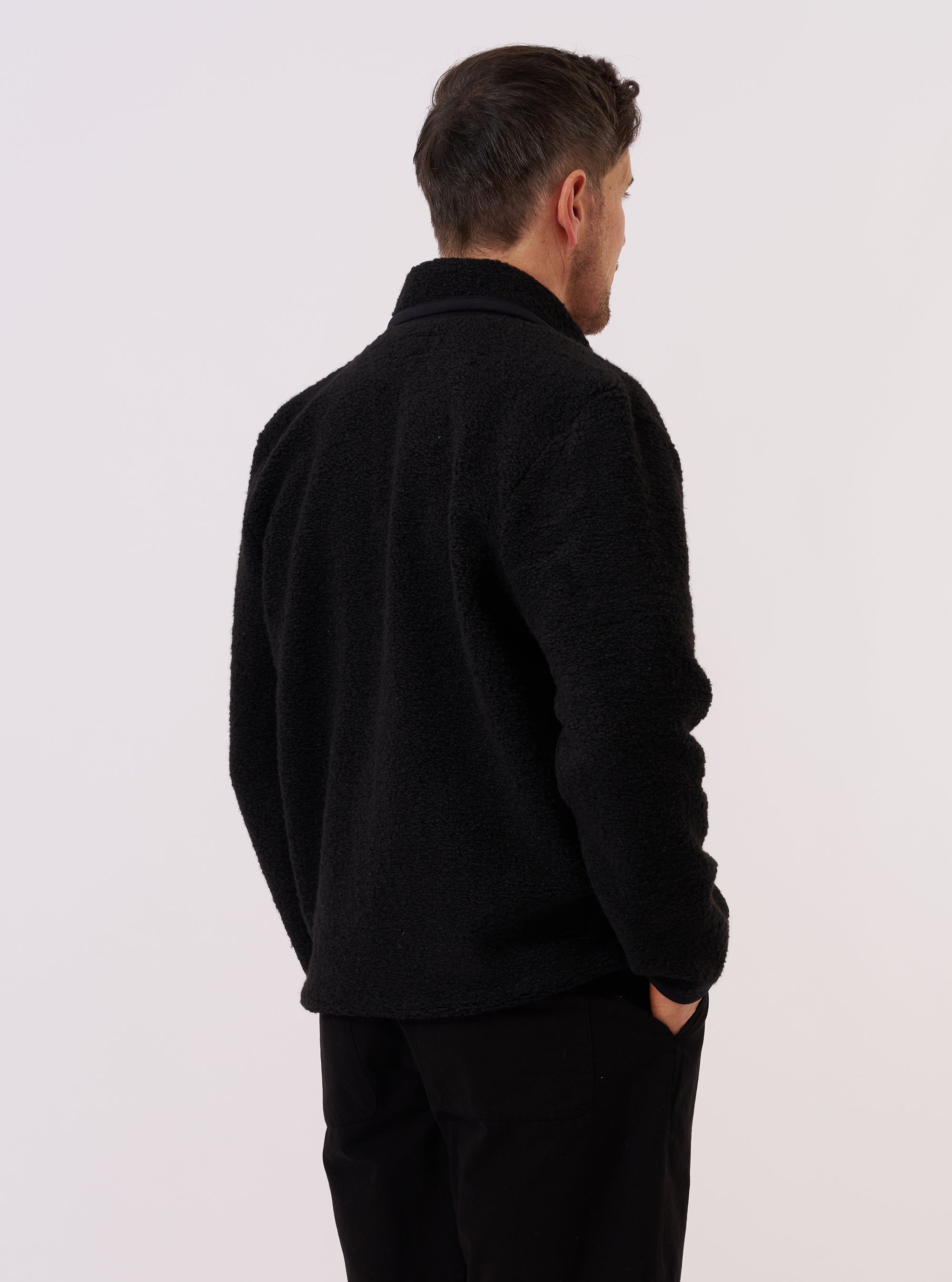 A model wears the Black Alvar Fleece Lancaster Jacket, shown from the rear against a plain white background. The jacket features a rounded shawl collar, a straight hem, and a relaxed fit. The black fabric has a distinct, curly fleece texture. No mannequin is present in the image.