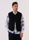 A model wears the Black Alvar Fleece Lancaster Gilet over a light-colored long-sleeve top, standing against a plain white background. The gilet features a deep V-neckline, a central button-down closure, and two large patch pockets at the waist. The black fabric has a soft, textured fleece appearance with tonal trim along the edges and pockets.