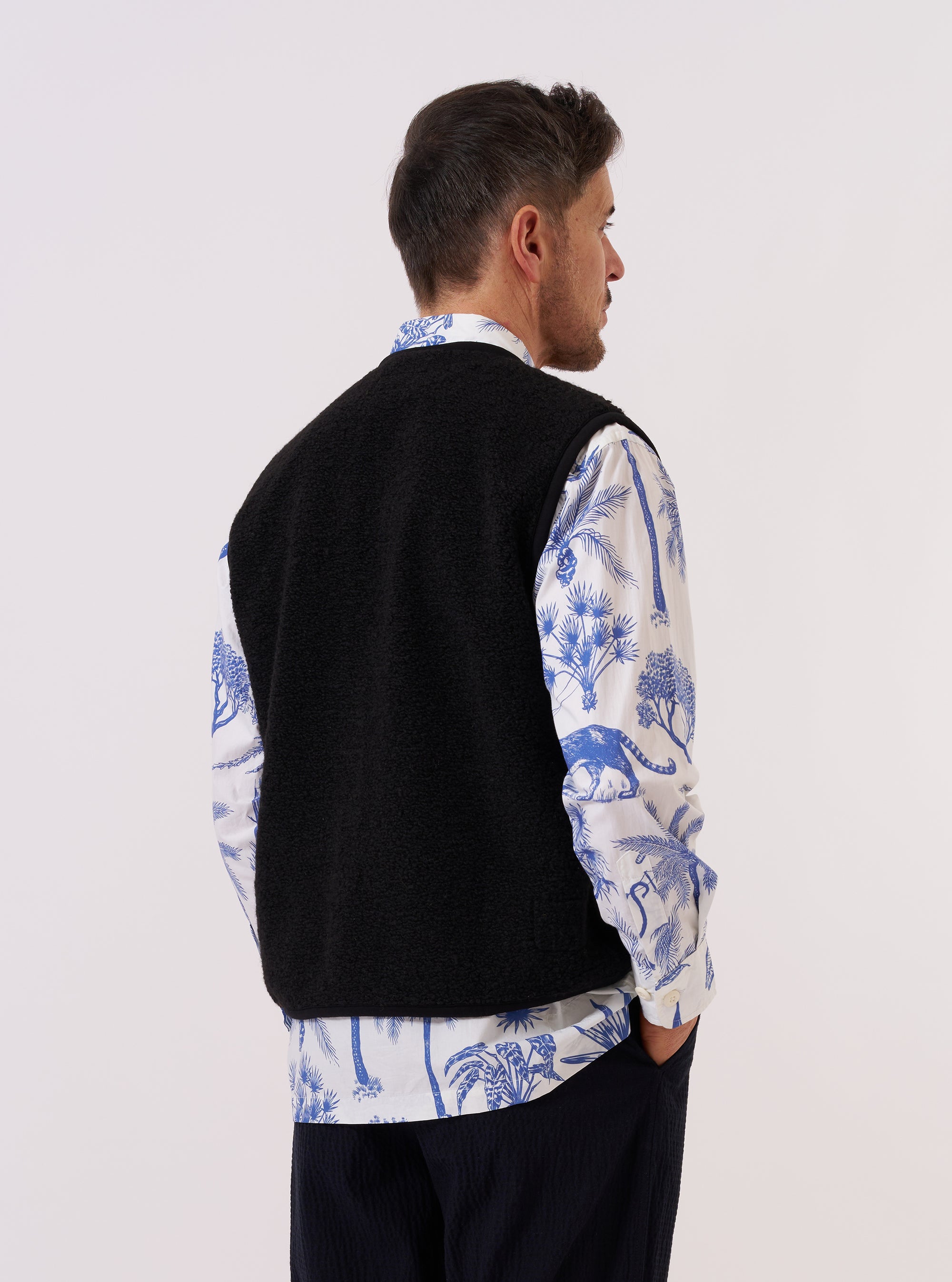 A model is wearing the Black Alvar Fleece Lancaster Gilet over a light-colored long-sleeve top, shown from the back against a plain white background. The gilet features a soft, curly fleece texture in black and a relaxed fit with a straight hem. The sleeveless design and rounded shoulders are prominent in this rear-view image. No mannequin is present.
