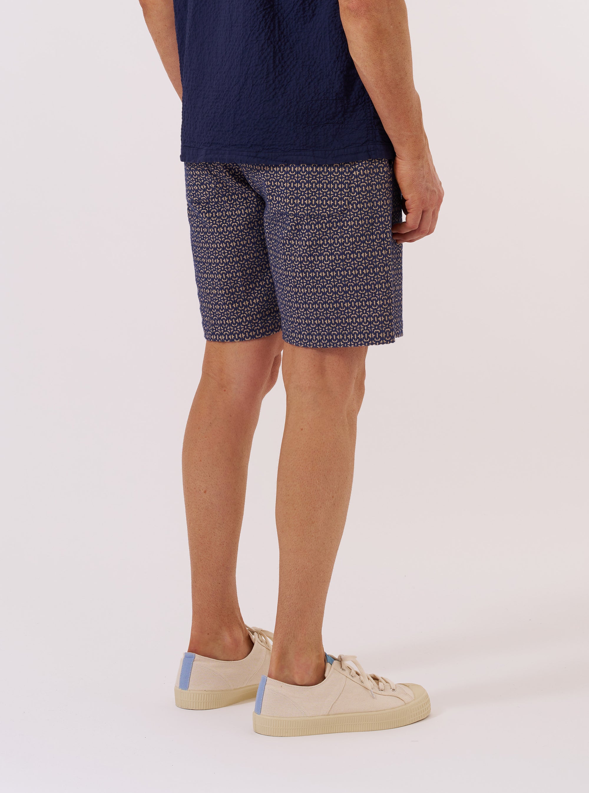 A model wears the Navy Konos Cotton Simple Short against a plain white background. The shorts feature a navy blue cotton fabric with a white repeating pattern throughout. The model is shown from the waist down, highlighting the garment's relaxed fit, elasticated waistband, and mid-thigh length.