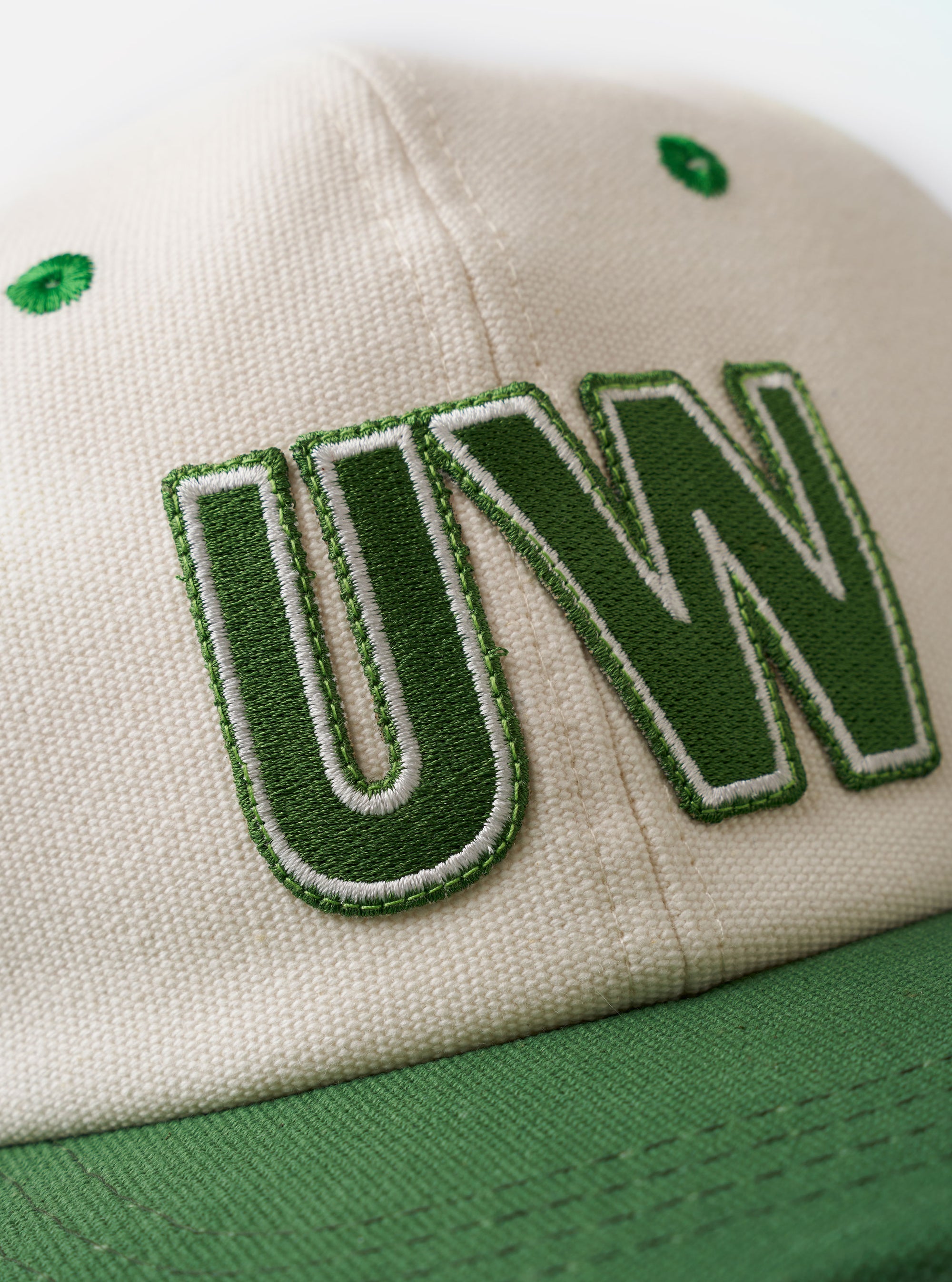 The Ecru/Green Canvas Baseball Hat is displayed as a flat lay against a plain white background. The cap features a light ecru crown with a contrasting green peak. It is constructed from a durable canvas fabric and includes tonal stitching and a curved brim.