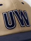 The Sand/Navy Canvas Baseball Hat is shown as a flat lay against a plain white background. The hat features a sand-colored canvas crown contrasted with a navy blue brim and a matching navy top button. It is constructed with six panels, tonal embroidered eyelets, and a curved peak.