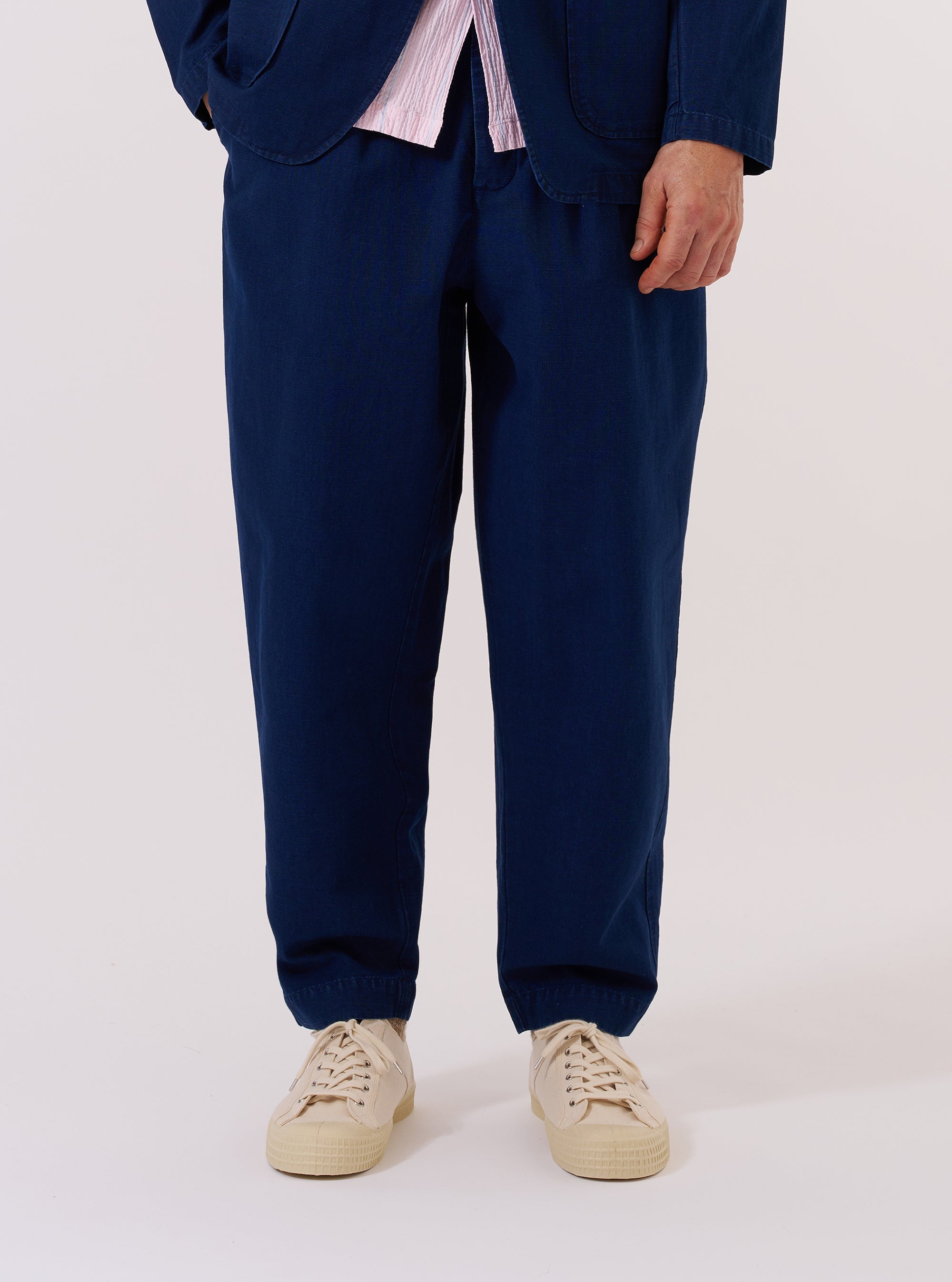 A model wears the Indigo Kimstop Pleated Track Pant while standing against a plain white background. The image shows a front view of the trousers, highlighting the loose fit, tapered leg, and single pleat front. The elasticated waistband and drawstring are visible, along with two front pockets. The indigo-colored fabric features a subtle ripstop texture. The model is wearing the trousers with a light-colored top and white shoes.