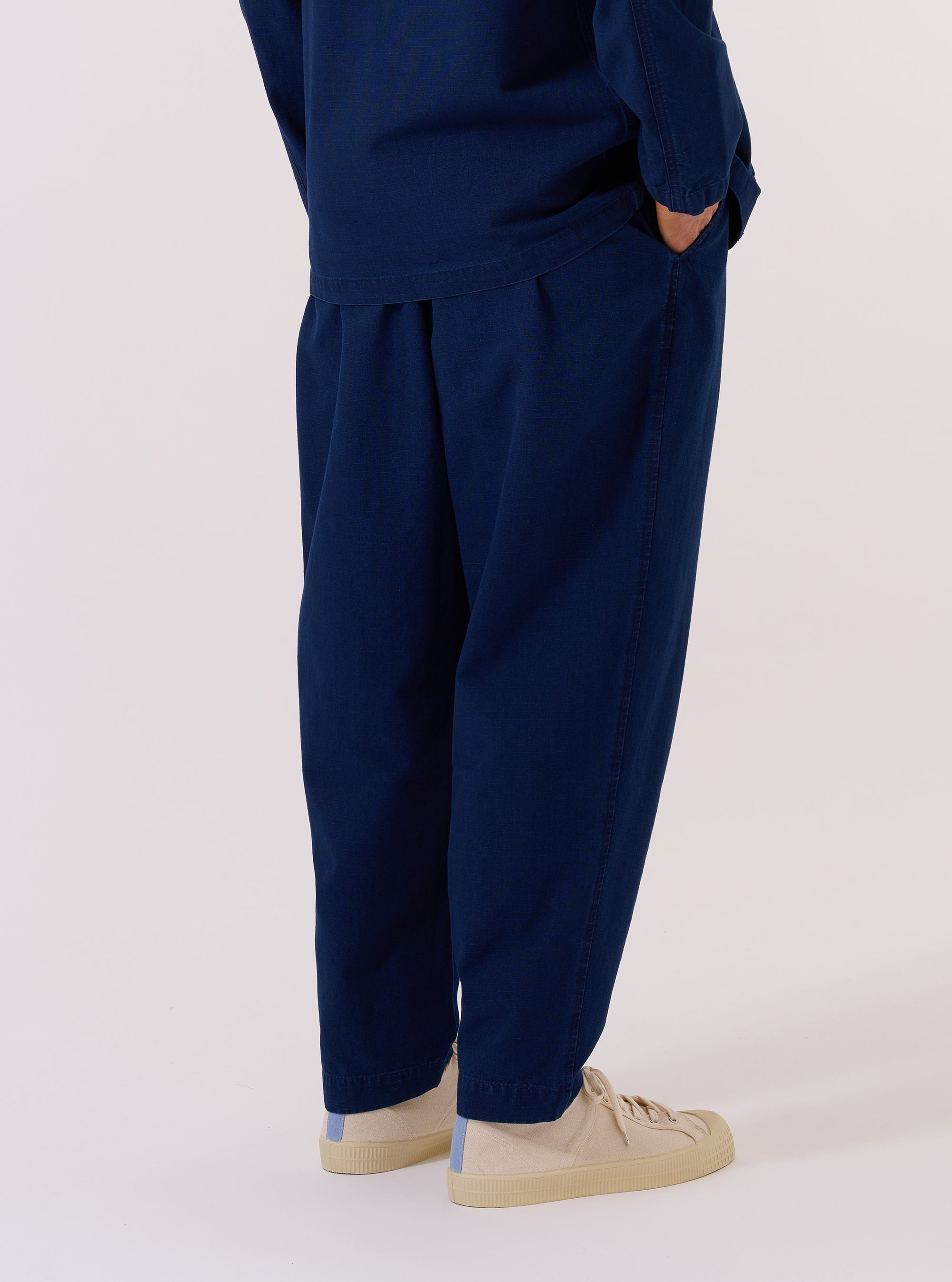 The Indigo Kimstop Pleated Track Pant is worn by a model against a plain white background, shown from the waist down from a rear three-quarter angle. The image highlights the relaxed, tapered fit of the trousers and the deep indigo-dyed ripstop fabric. One rear pocket with a buttoned flap is visible, along with the elasticated waistband and clean hem. No mannequin is present in the image.