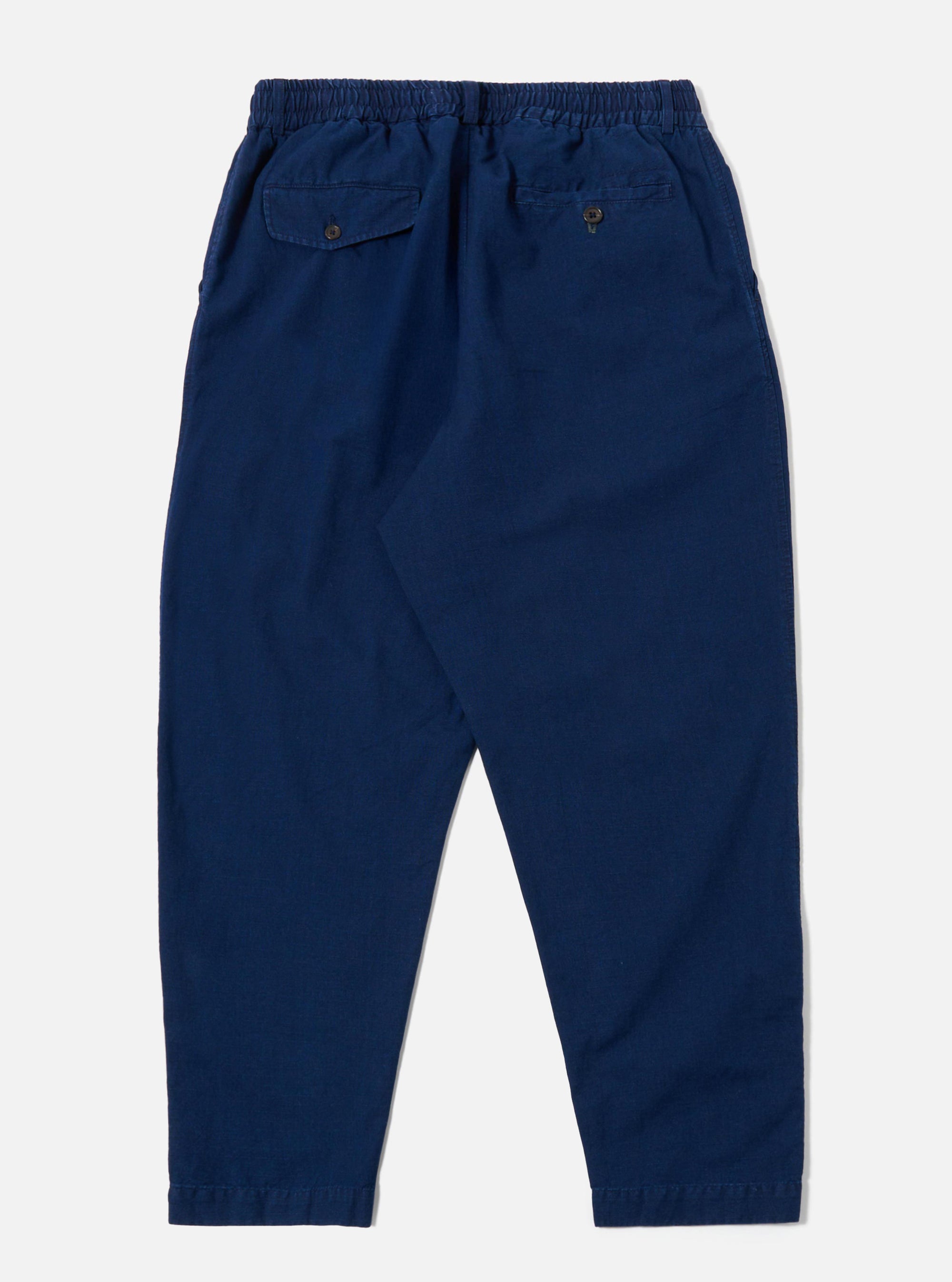 The Indigo Kimstop Pleated Track Pant is shown from the back as a flat lay against a plain white background. The trousers feature an elasticated waistband and two rear patch pockets. The garment is made from a dark indigo organic cotton fabric with a visible textured weave.