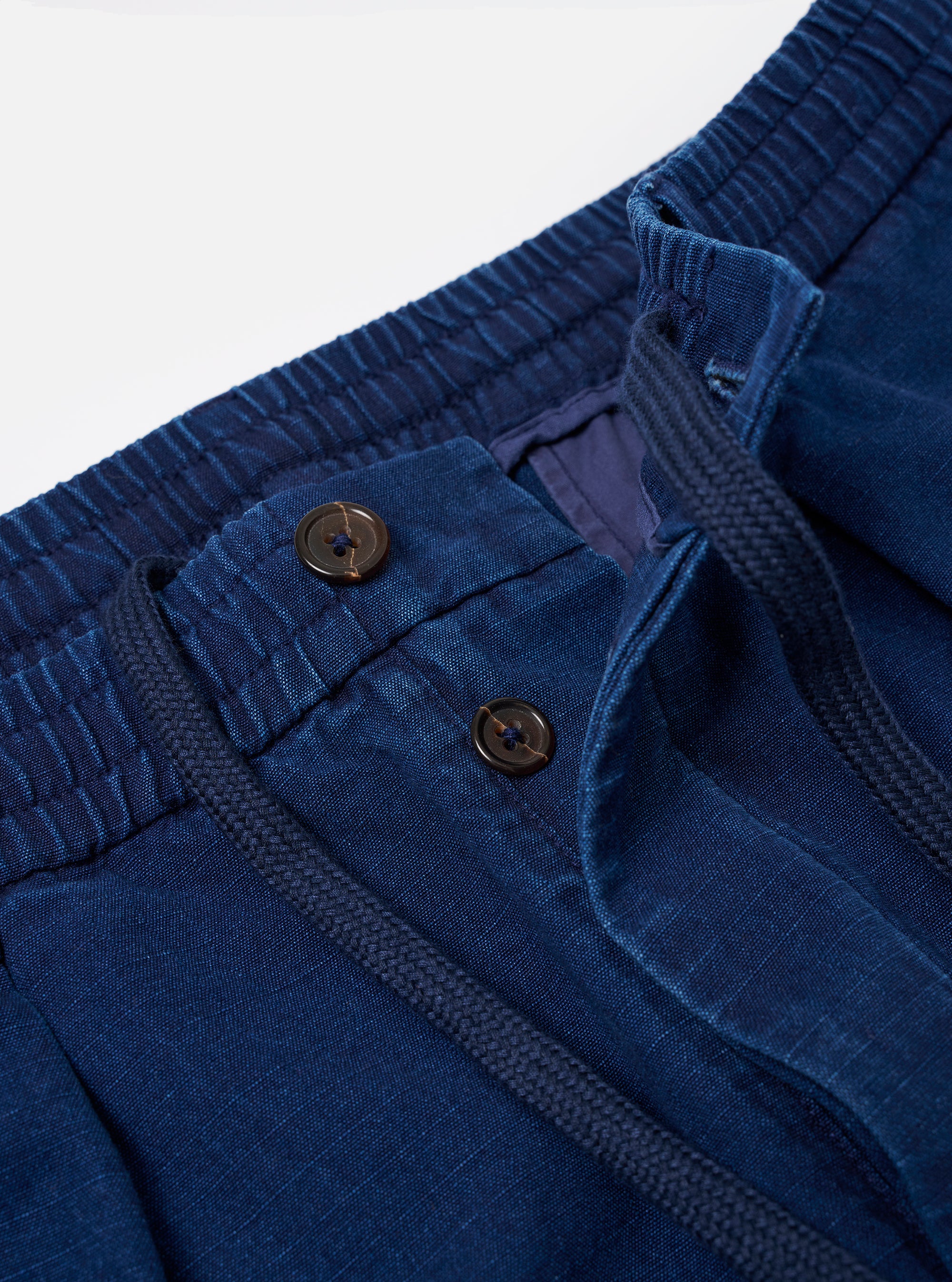 The Indigo Kimstop Pleated Track Pant is shown in a close-up detail shot against a plain white background. The image focuses on the top section of the trousers, highlighting the elasticated waistband with an internal drawstring, a single pleat on the front, and the deep indigo blue color of the organic cotton fabric.