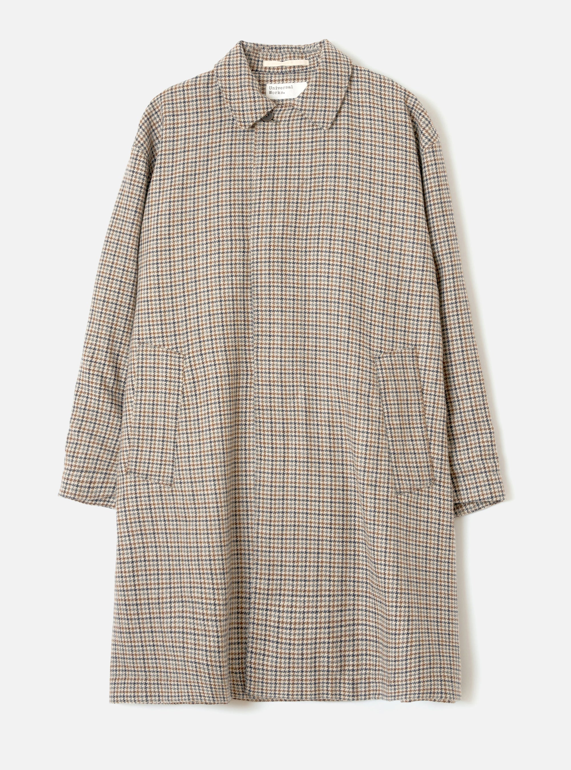 Taupe dogtooth check Stan Coat by Universal Works, laid flat on a light grey seamless backdrop. The longline coat features a soft point collar, concealed front placket, long sleeves with plain cuffs, two large patch pockets at the hip, and a relaxed oversized silhouette. The woven fabric shows a tan, navy and cream dogtooth pattern.