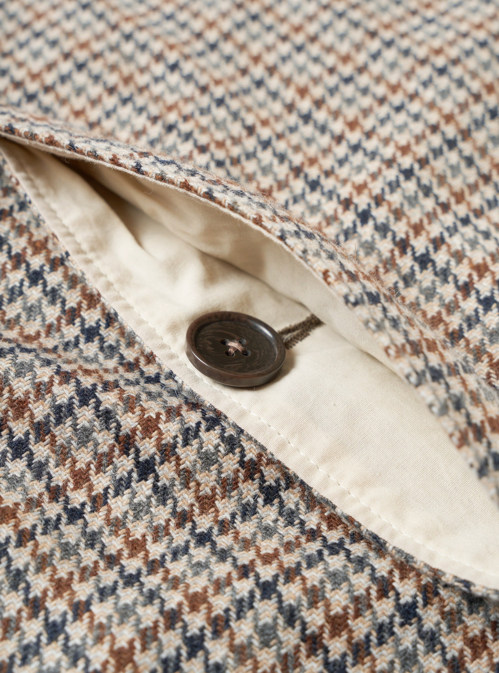 Close-up detail of the taupe dogtooth Stan Coat. A dark brown four-hole button sits on a cream woven lining visible through an open chest pocket. The outer fabric shows a multicolour dogtooth weave in taupe, brown, navy and grey. Shot on a plain white studio backdrop.