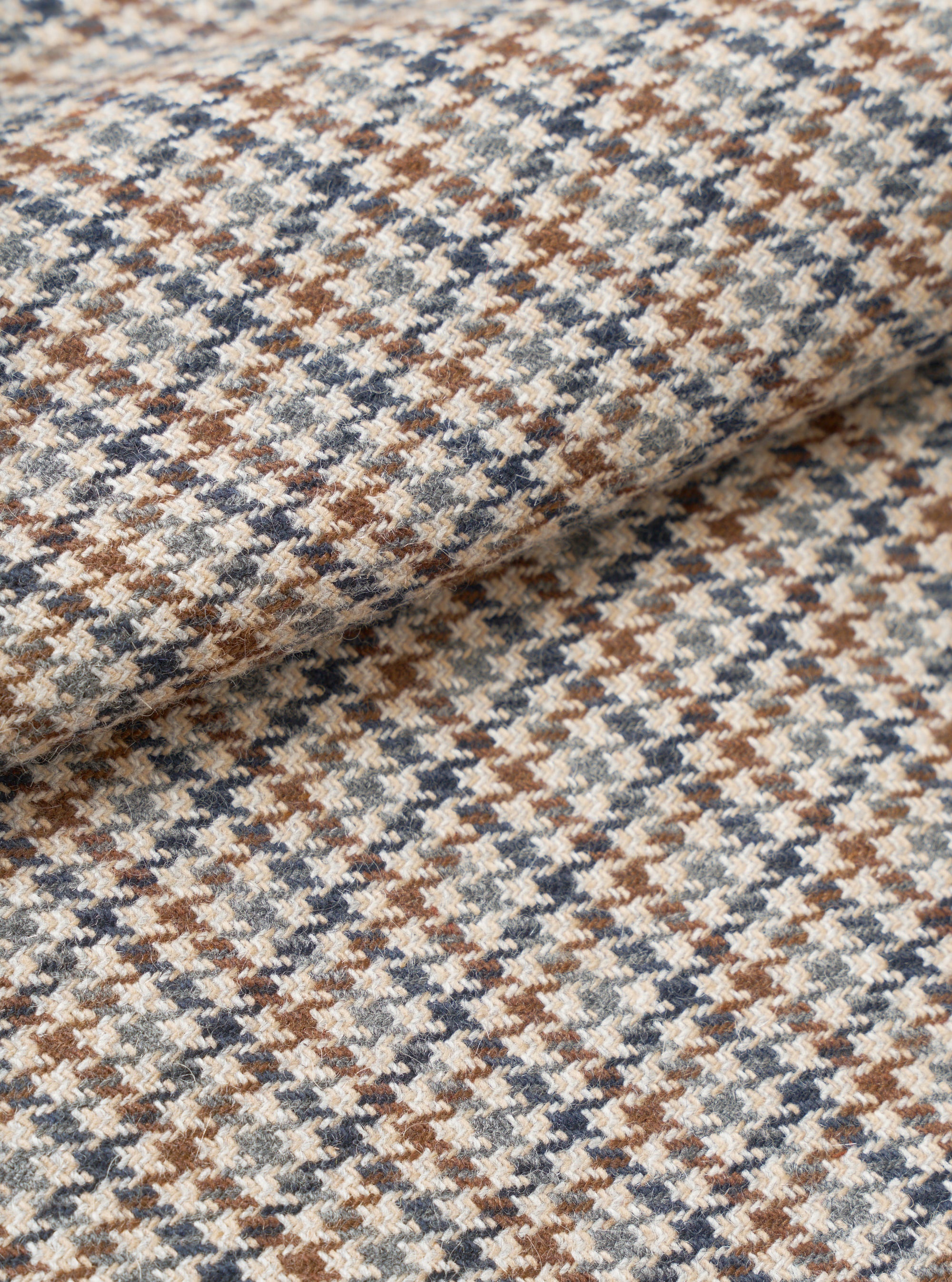 Close-up fabric detail of the Lester Dogtooth Franck Jacket in taupe, showing a woven wool-effect dogtooth pattern in cream, tan brown and slate blue. The fabric is folded to reveal texture and weight. No model or mannequin is present.