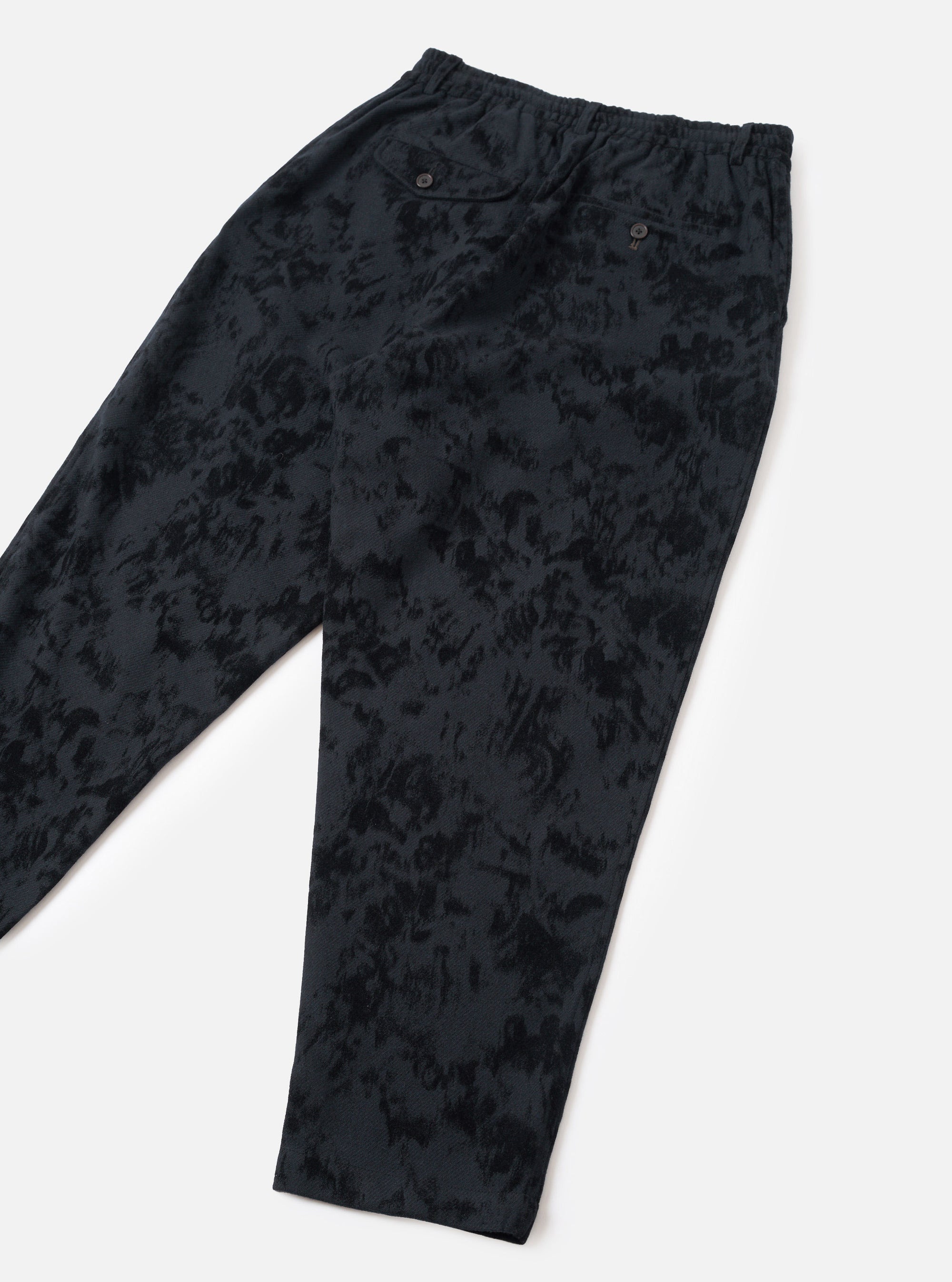 Flat lay of black wool mix track pants on a light grey seamless backdrop. The fabric features a tonal abstract jacquard pattern in varying depths of black. An elasticated waistband sits at the top with two small dark buttons visible on the front. The tapered legs are shown at full length with no cuffs.