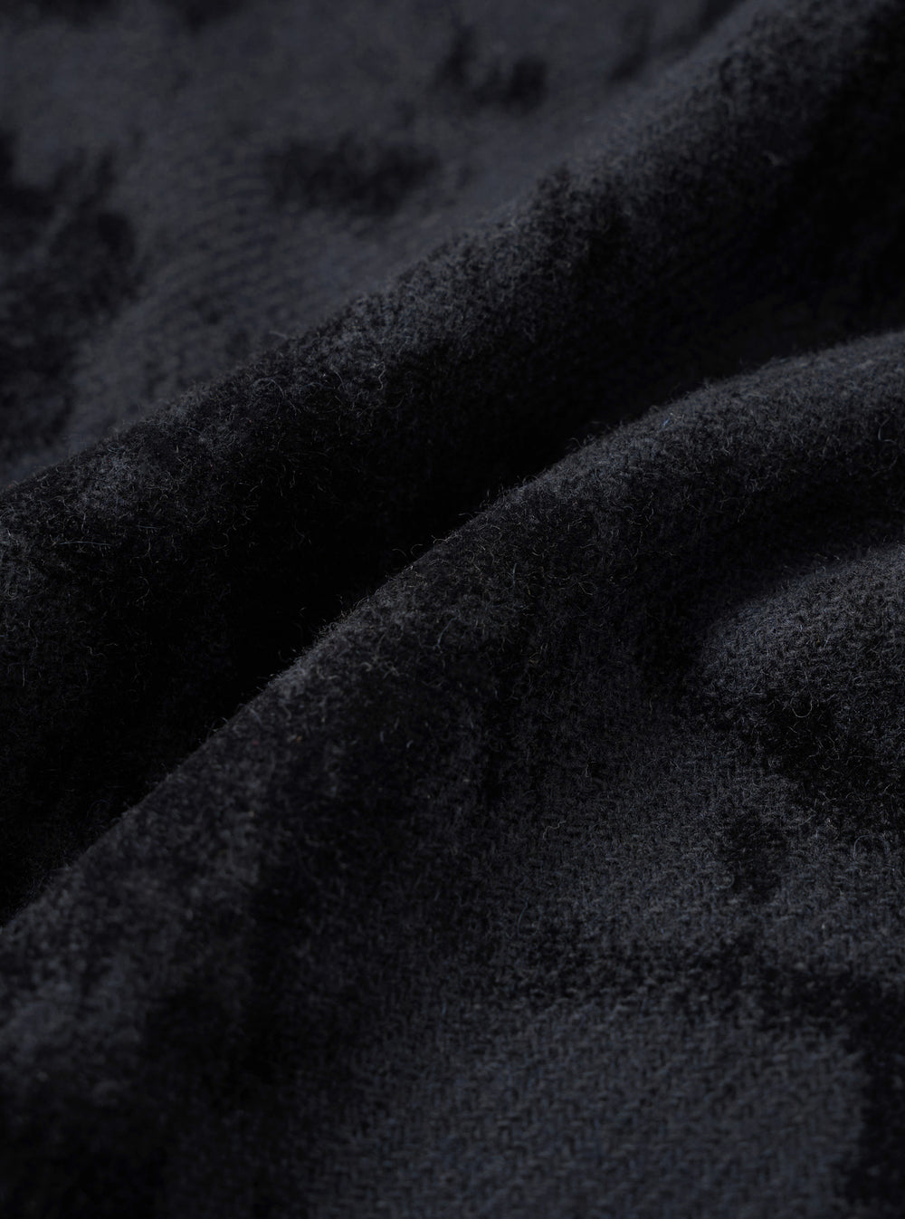 Close-up detail of the black wool mix fabric used in the pleated track pant. The surface has a dense, lightly boiled or brushed texture with fine fibres visible throughout. Deep folds in the fabric reveal soft drape and a matte finish with subtle tonal variation across the dark charcoal-black cloth.
